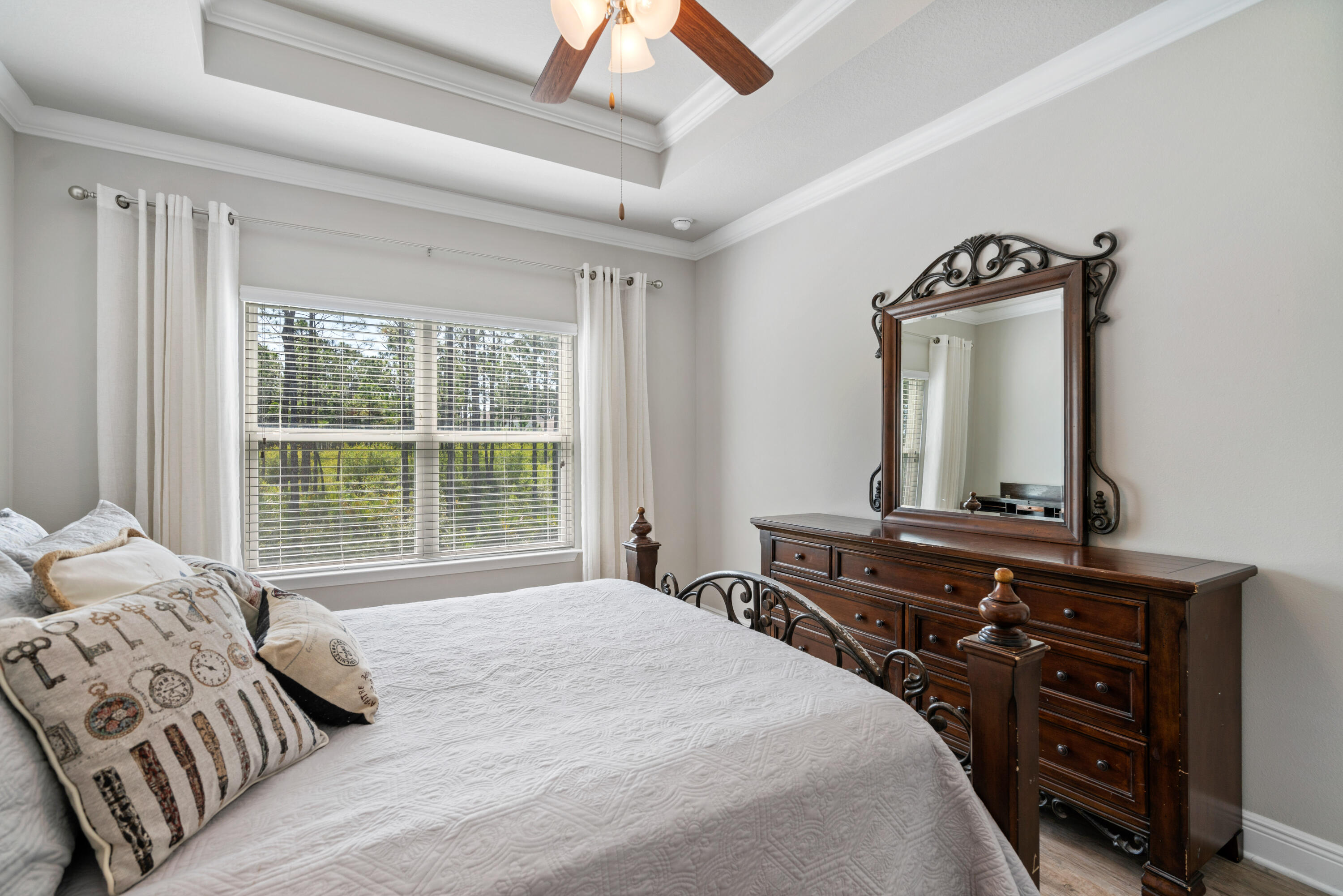Bedroom with a white quilted bed, carved wood furniture, and a large window overlooking trees.