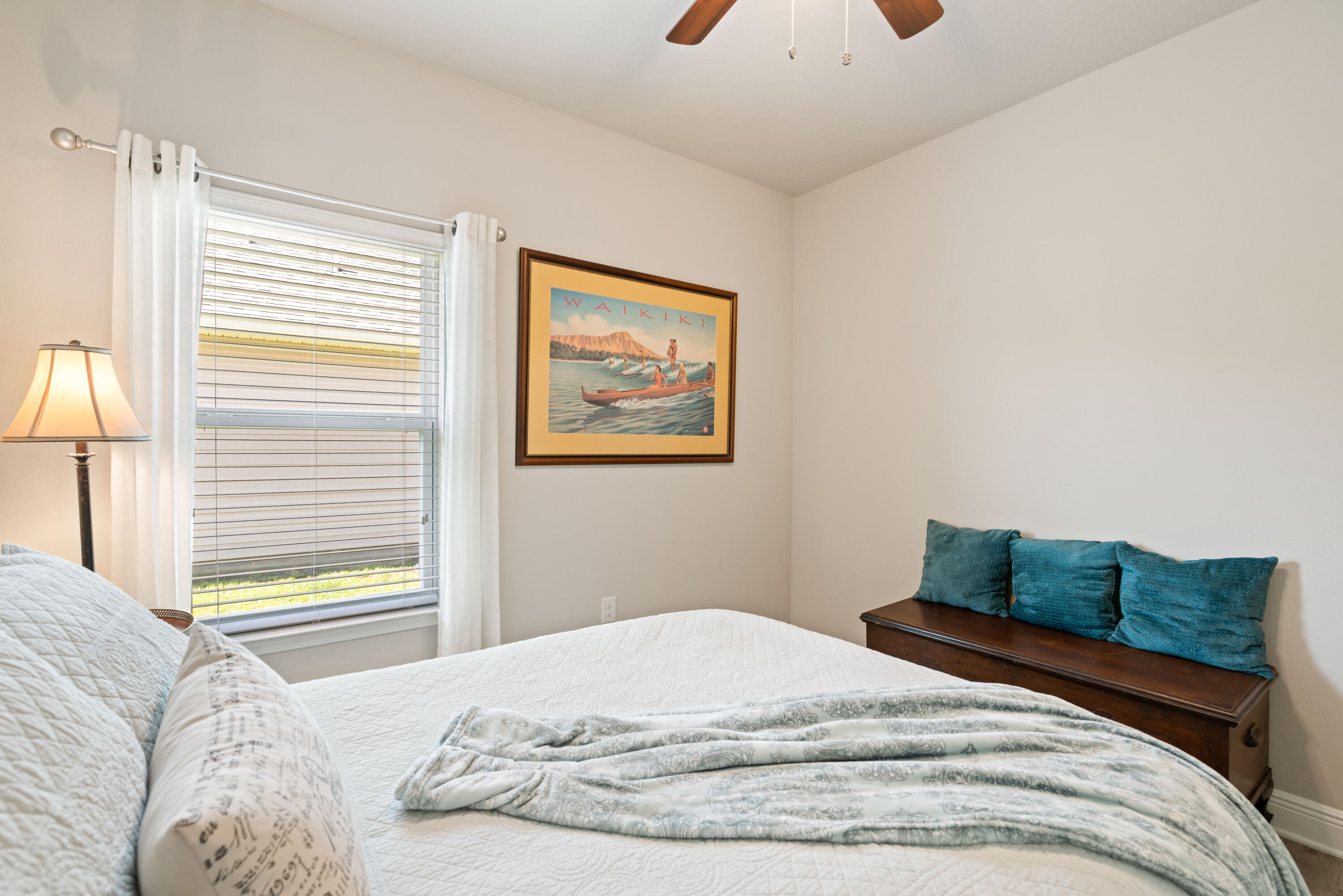 Bedroom with a white quilted bed, teal cushions on a wooden bench, and framed Waikiki artwork.