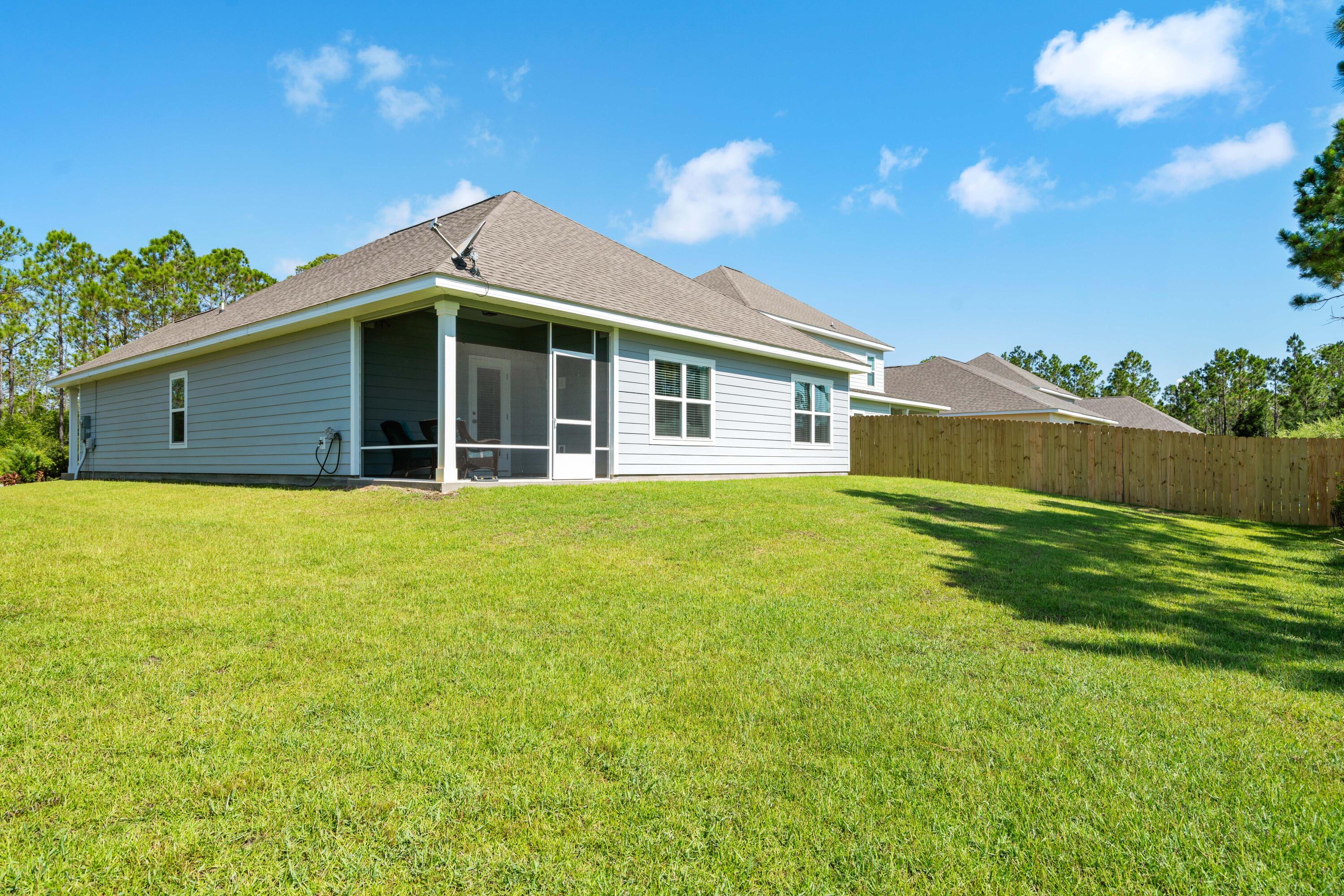 Rear of a light blue-gray house with a screened porch, wide lawn, and wooden privacy fence.