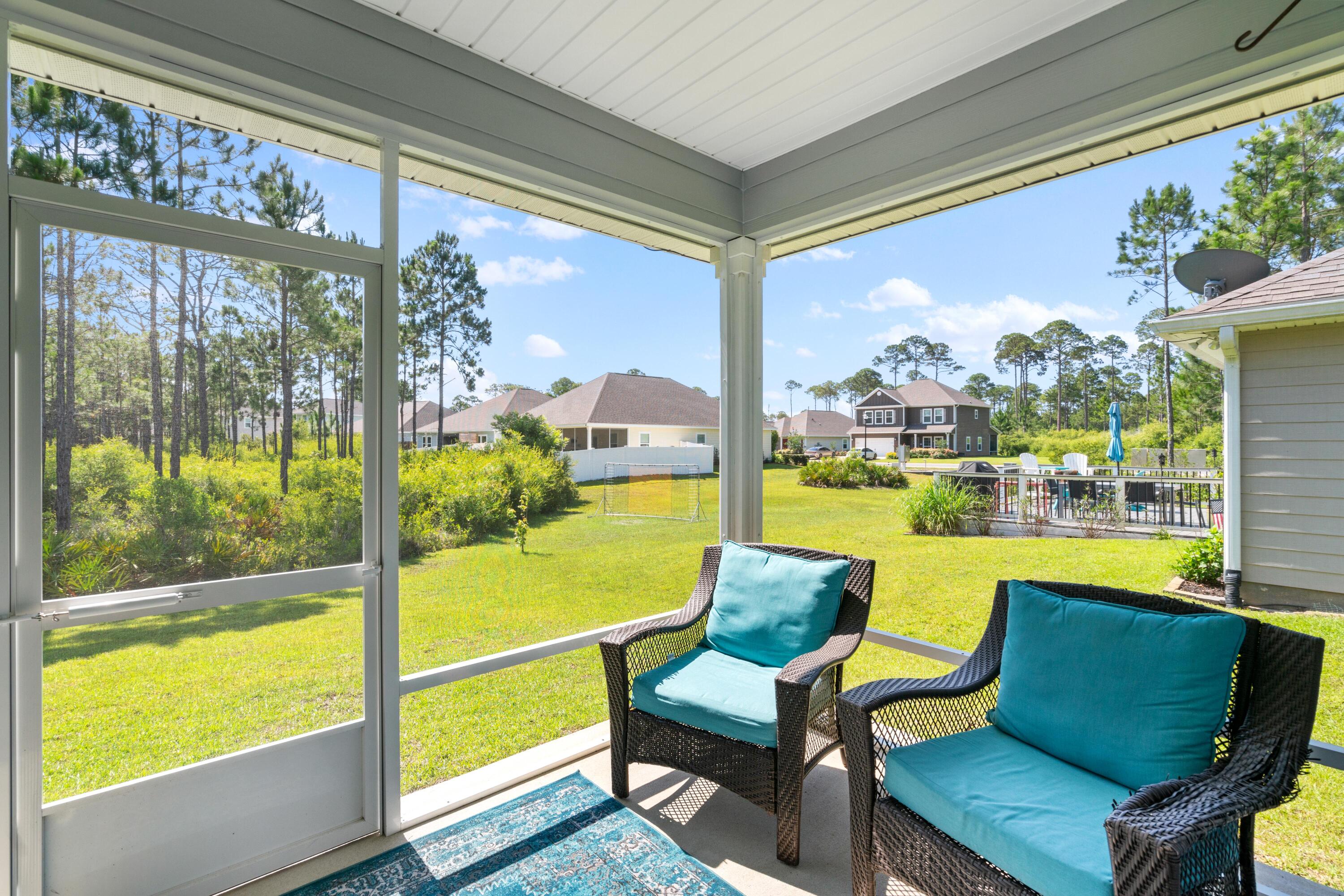 Screened porch with turquoise-cushioned wicker chairs overlooking a sunny lawn and pine trees.
