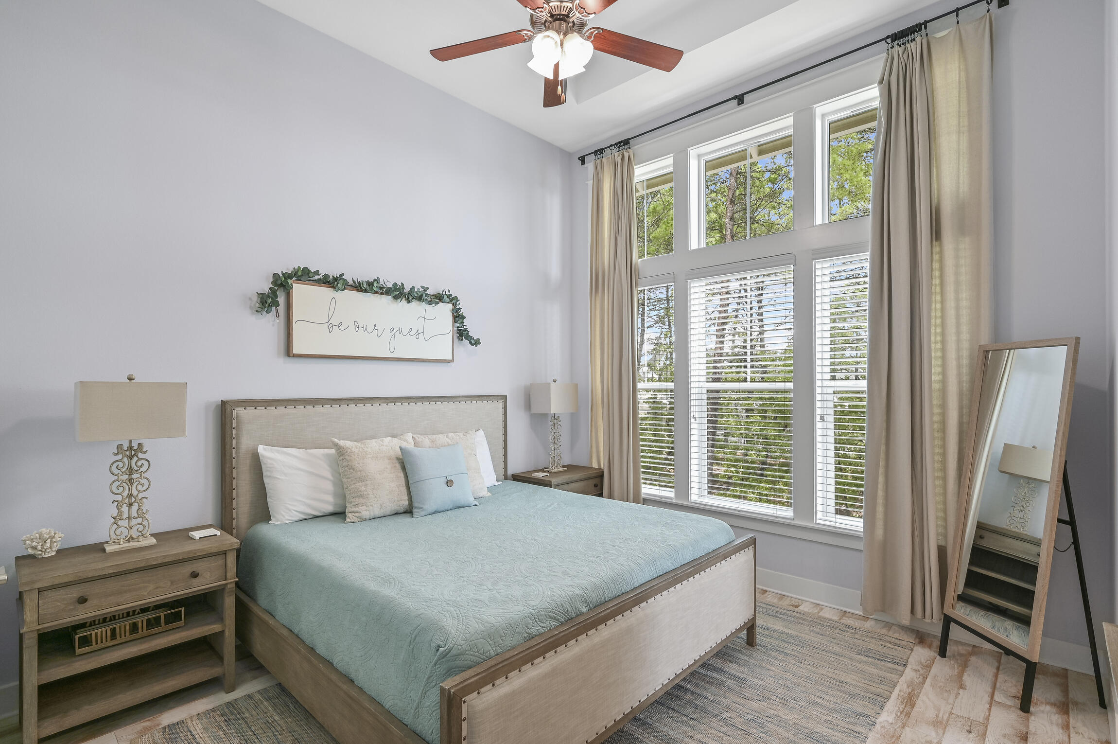 Staged bedroom with pale blue bedding, a tall bank of windows, and a framed “Be our guest” sign.