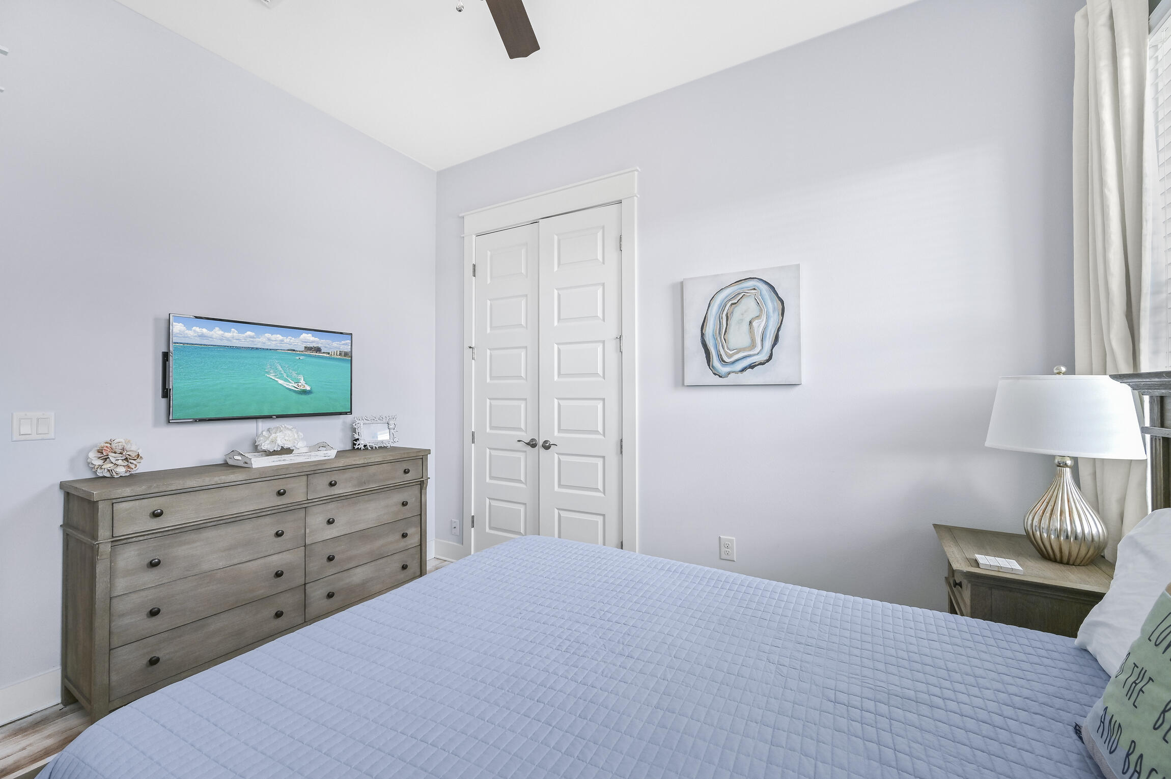 Furnished bedroom with a pale quilted bed, gray dresser, double doors, and television showing turquoise water.