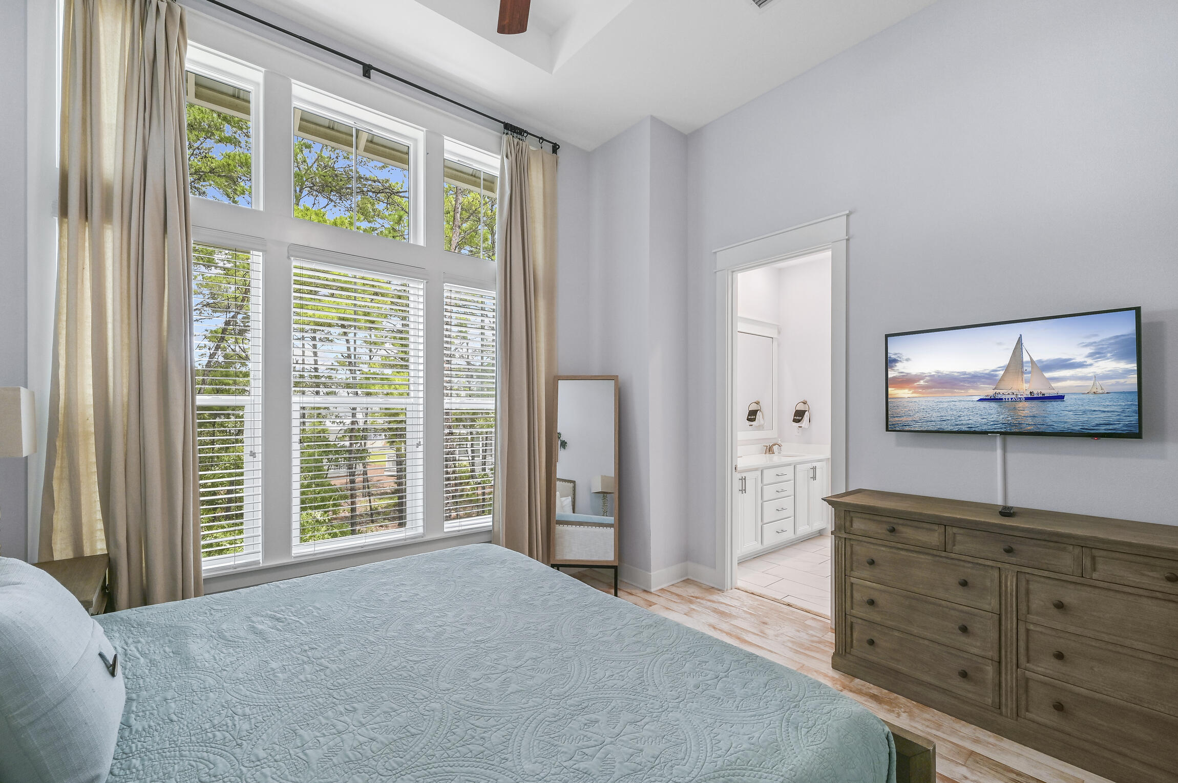 Bedroom with a pale blue quilt, tall curtained windows, and an adjoining bathroom vanity.