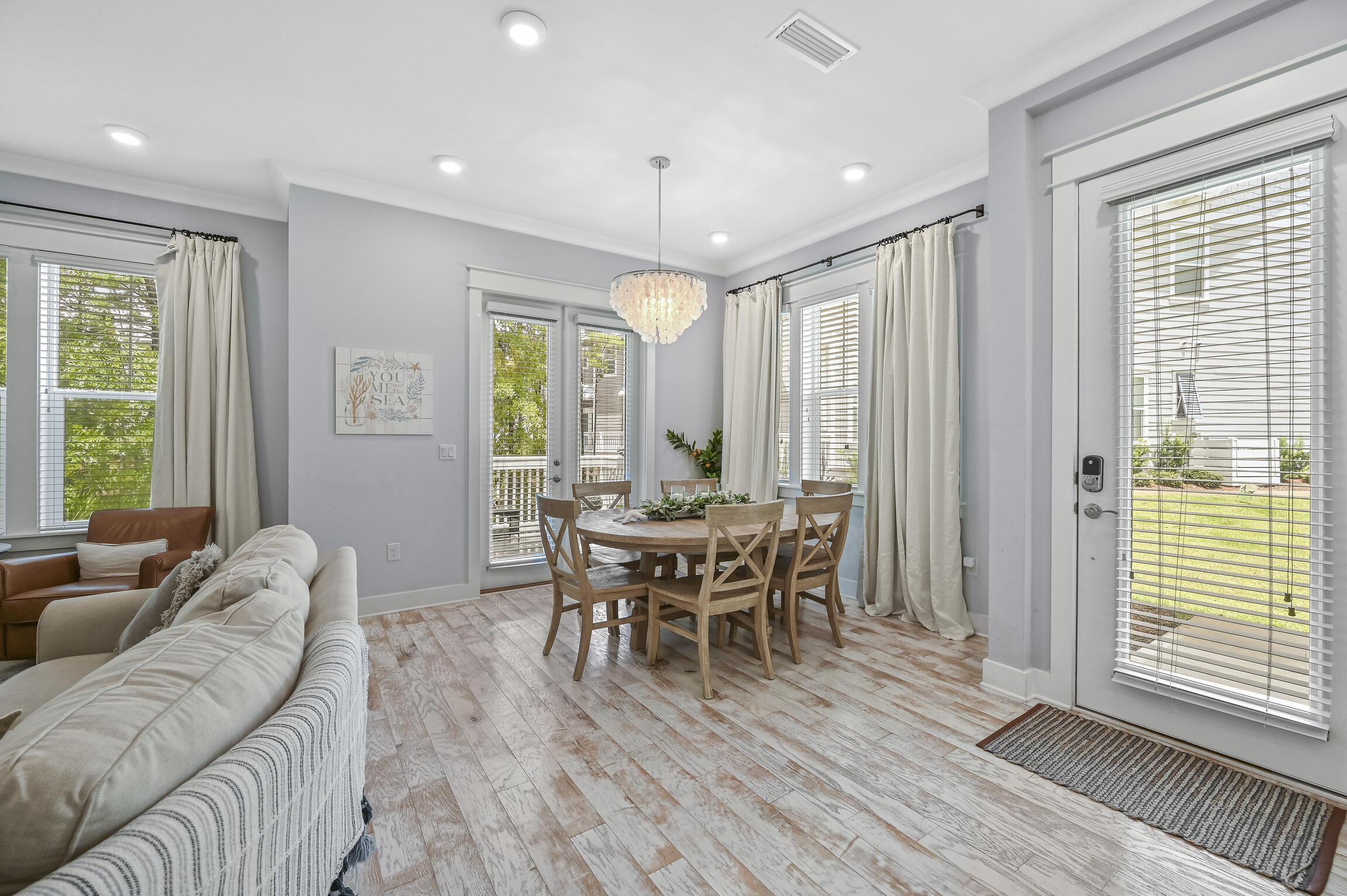 Open living and dining area with a round wooden table, shell chandelier, and multiple bright windows.