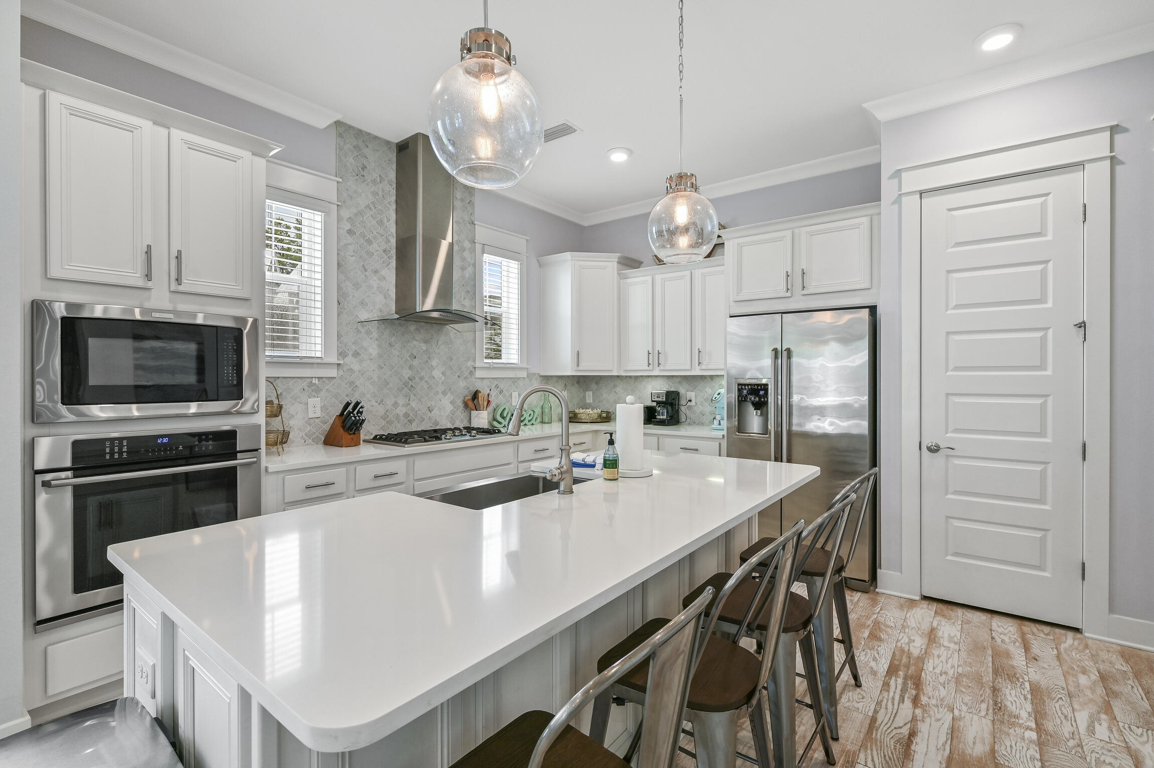 Modern white kitchen with a large island, stainless steel appliances, and glass pendant lights.