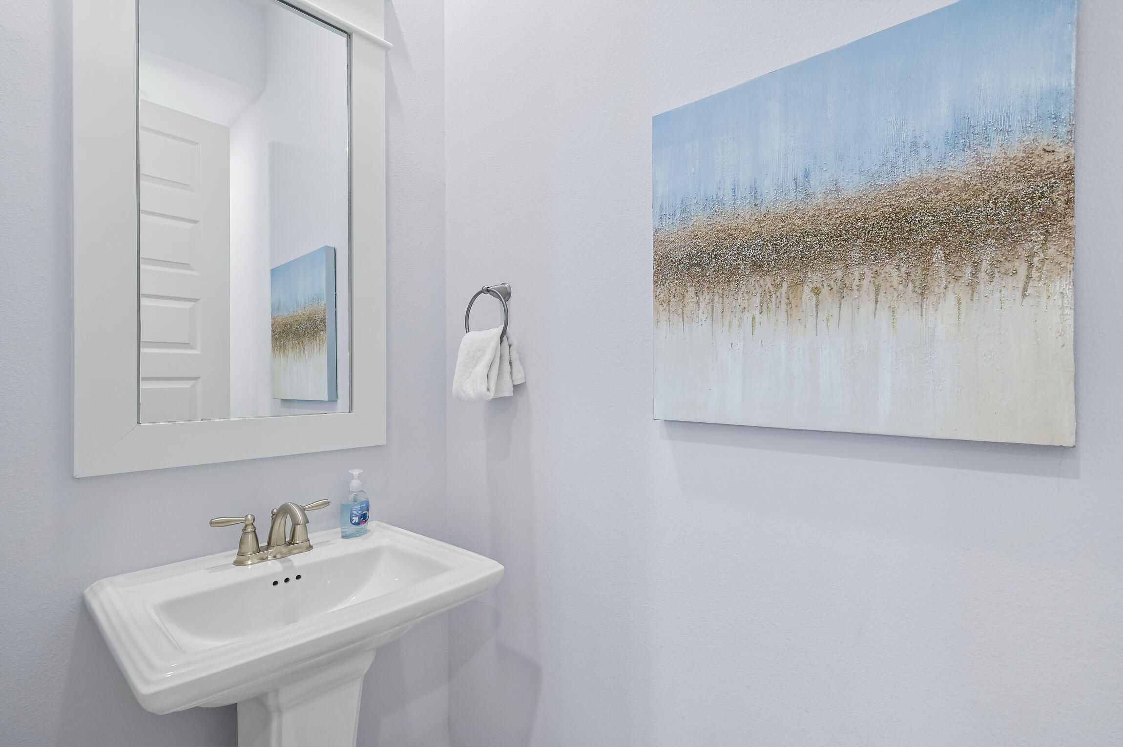 Bathroom pedestal sink beneath a framed mirror, beside a towel ring and abstract coastal-toned artwork.