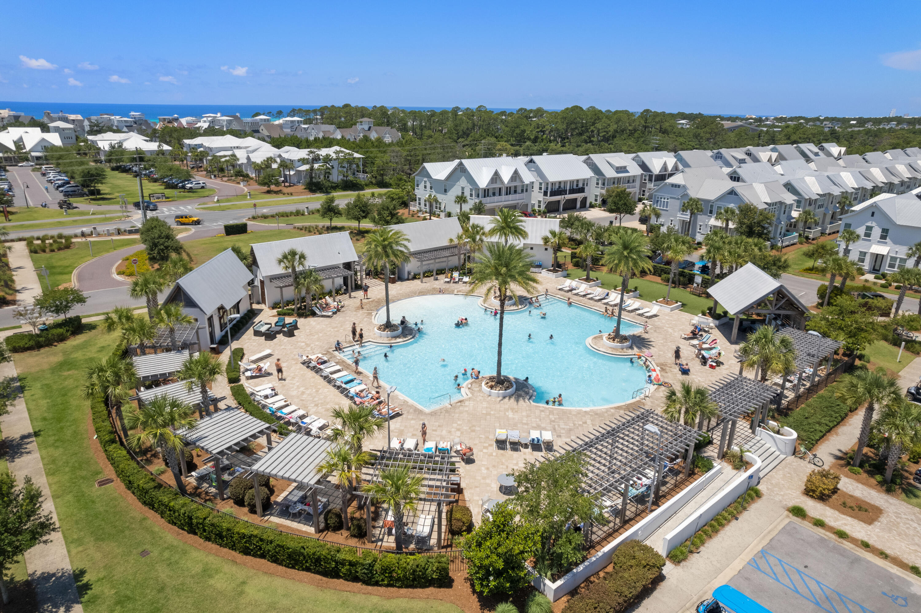 Aerial community pool with palm islands, surrounding townhomes, and ocean visible on the horizon.