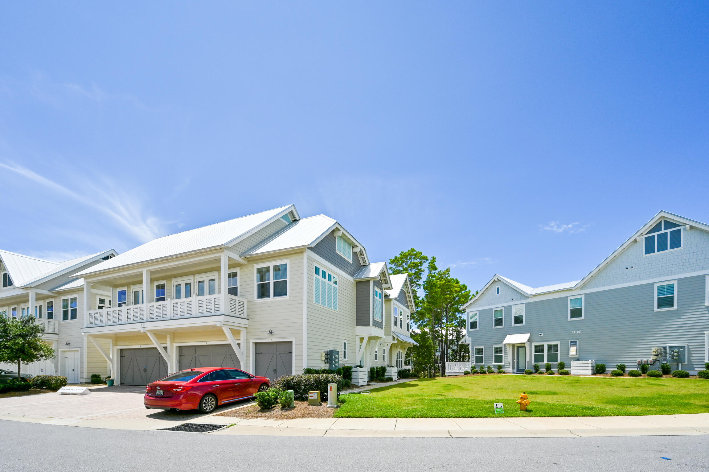Coastal-style homes with white metal roofs and balconies; a red car is parked beside the lawn.