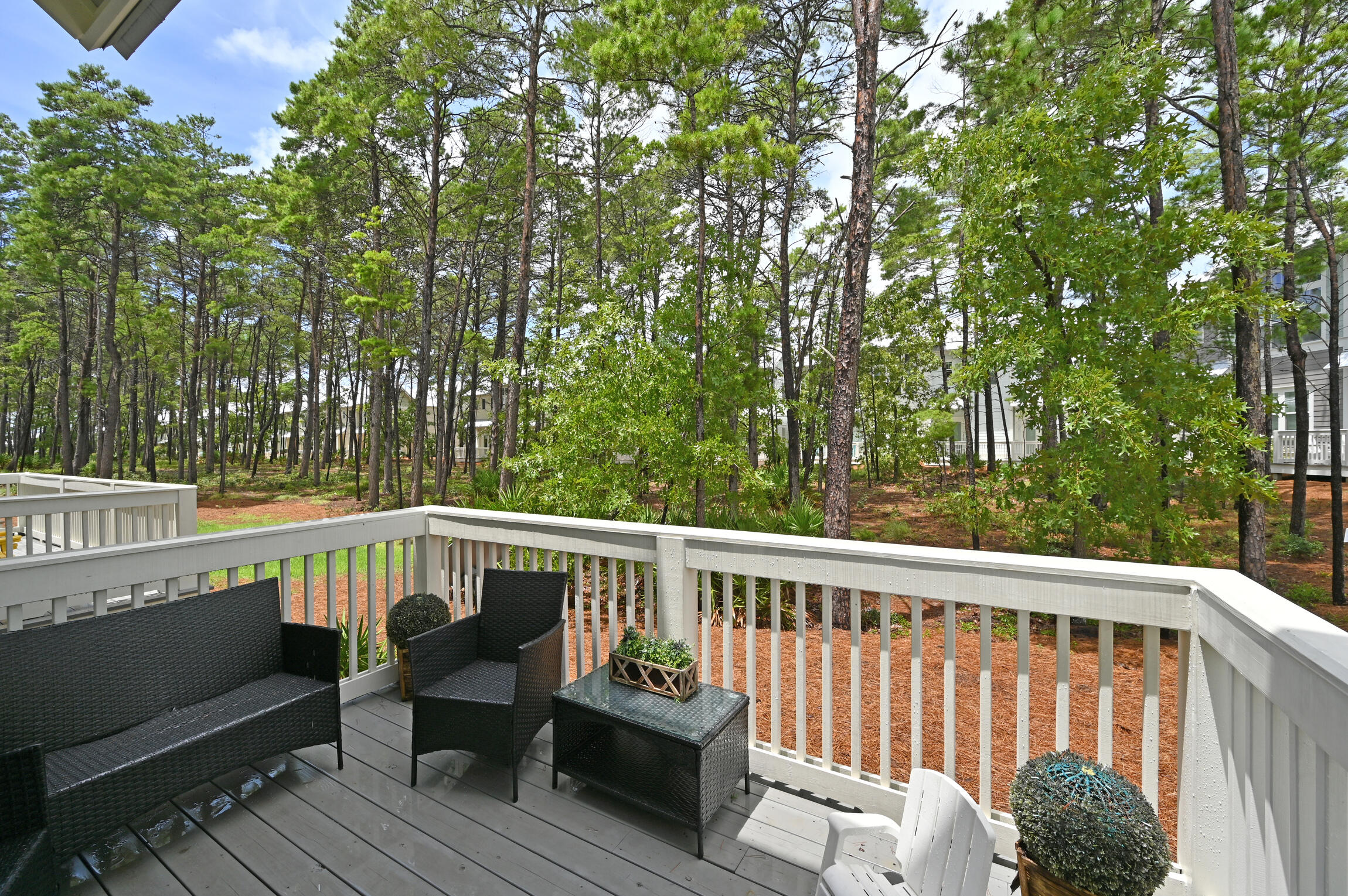 Furnished deck overlooking a wooded grove, with neighboring homes visible through the trees.