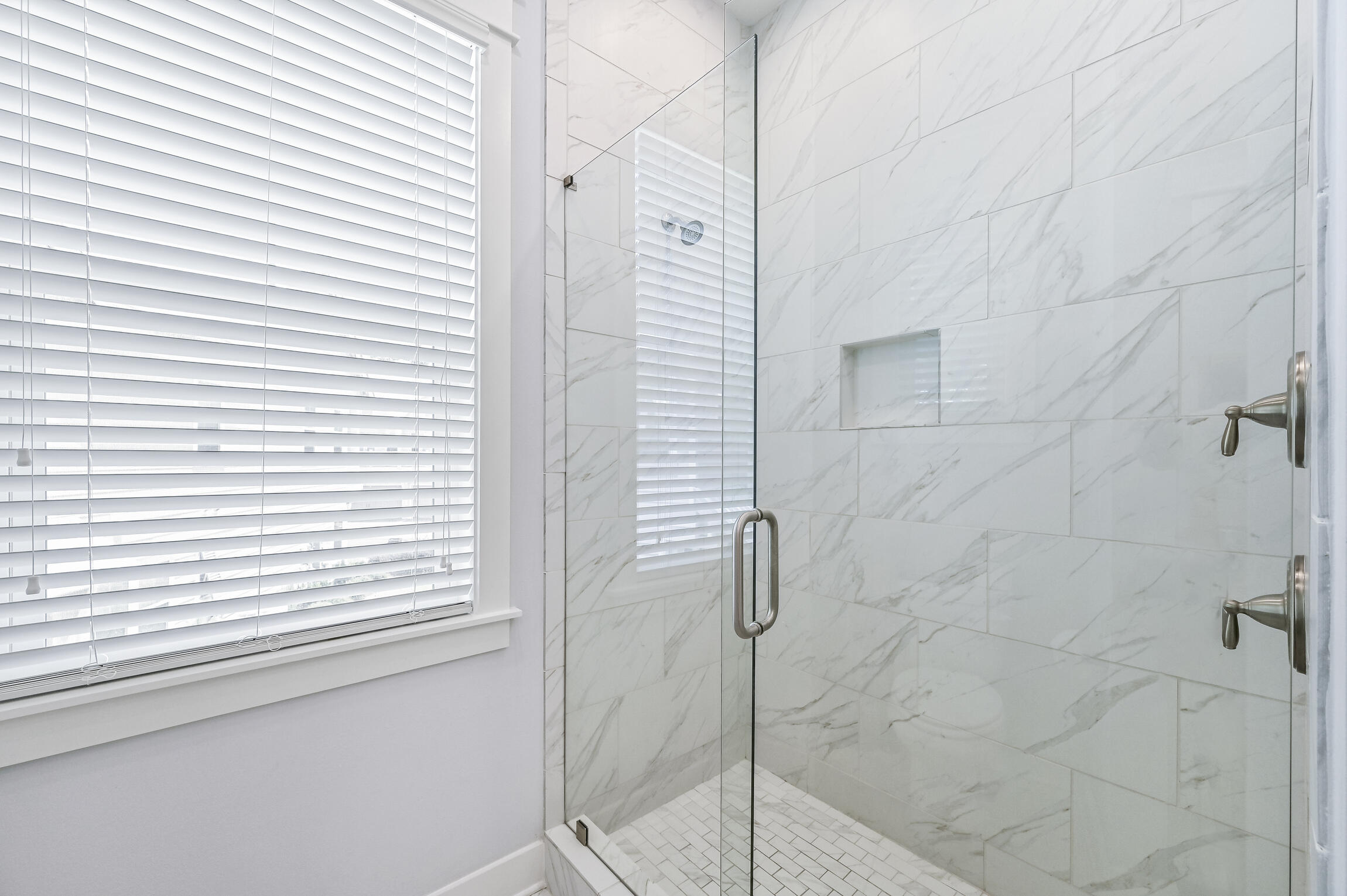 Glass-enclosed bathroom shower with marble-look tile, recessed niche, and a window with white blinds.