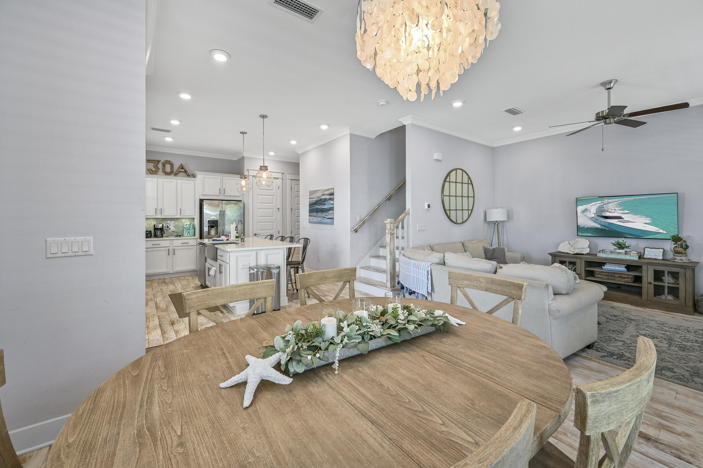 Open-plan living and dining area with a round wood table, white kitchen, pale sectional, and shell chandelier.