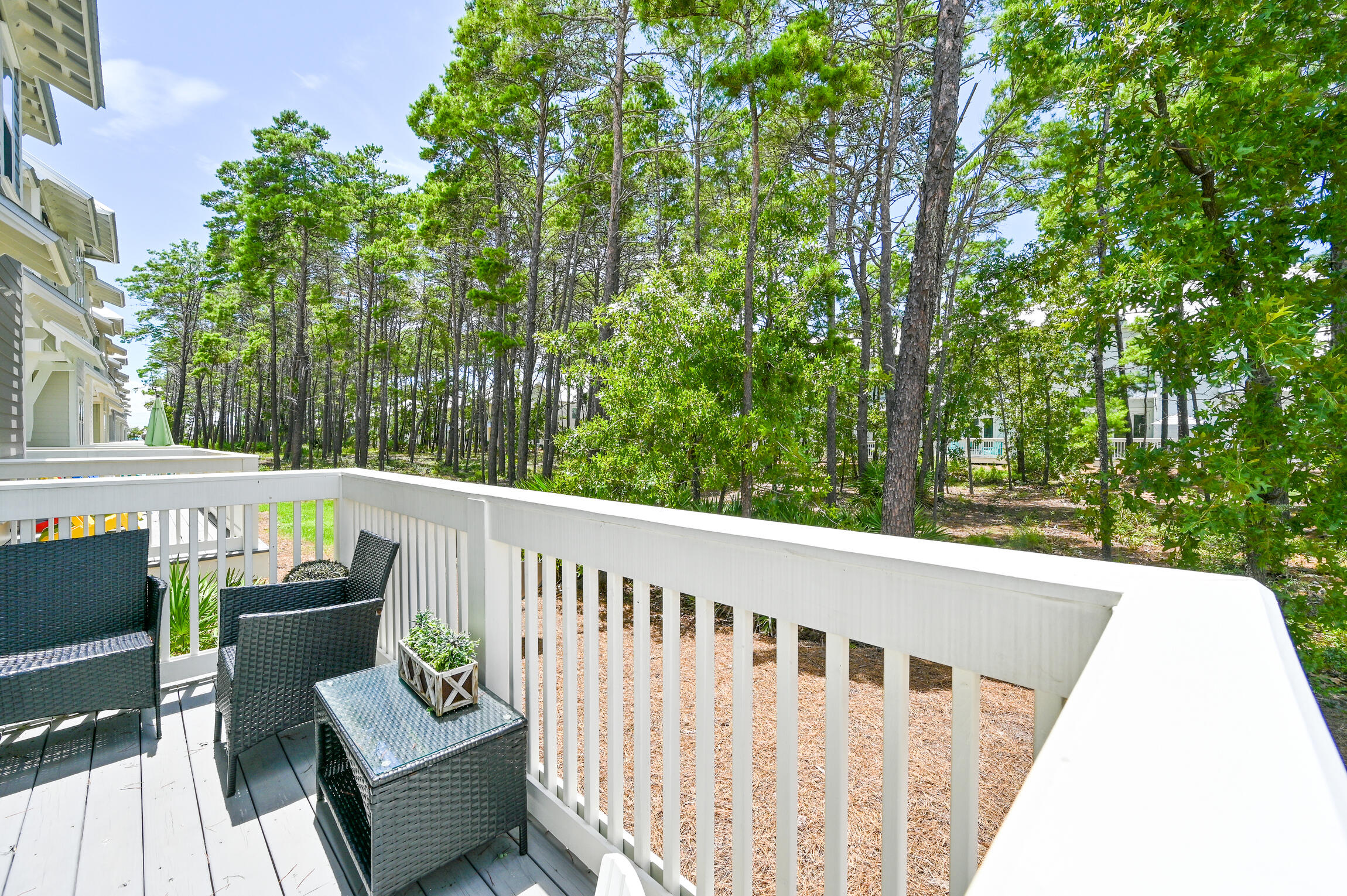 Furnished deck with white railings overlooking pine trees and neighboring homes.