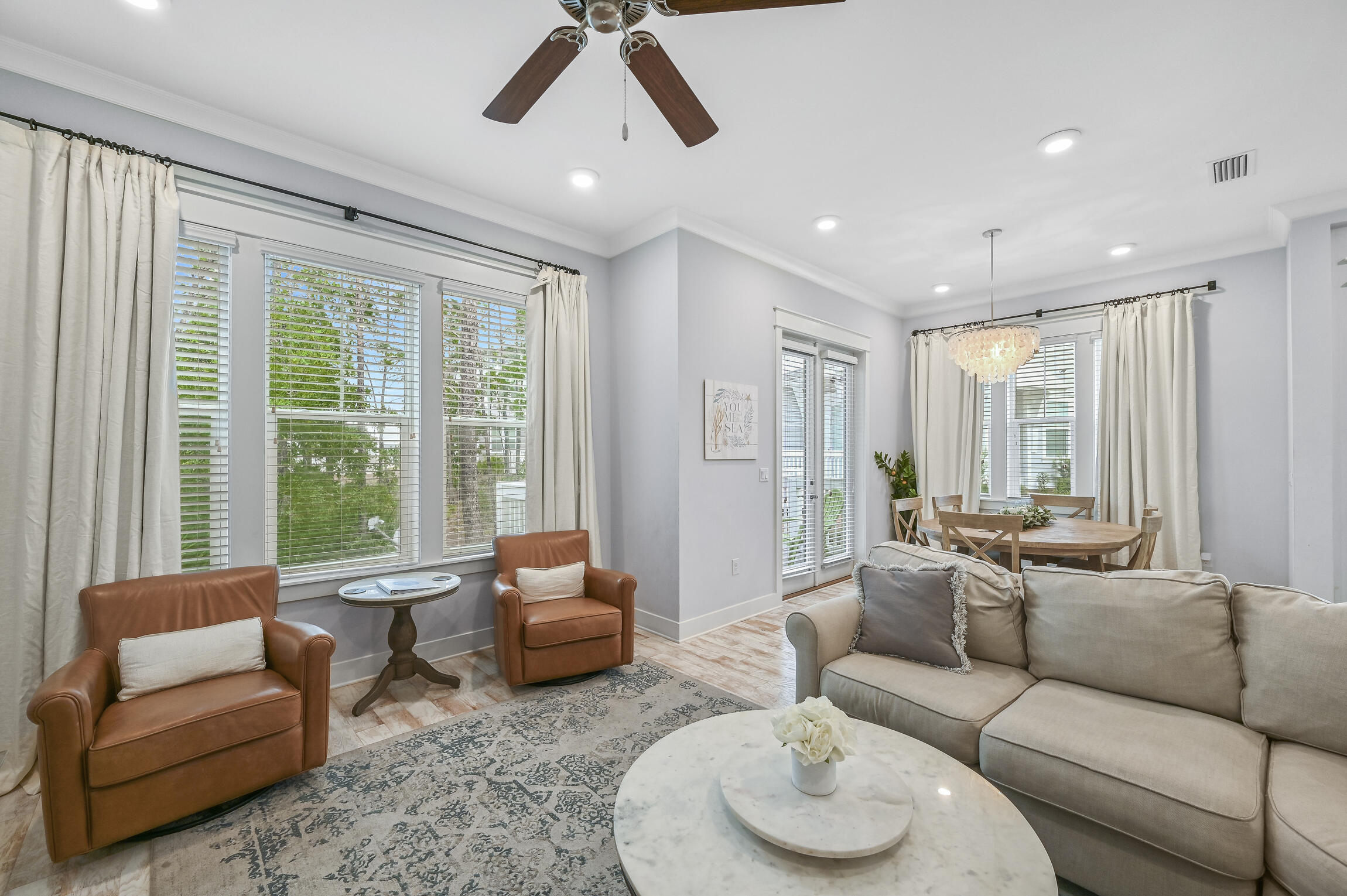 Open-plan living and dining room with a beige sofa, leather armchairs, and large curtained windows.