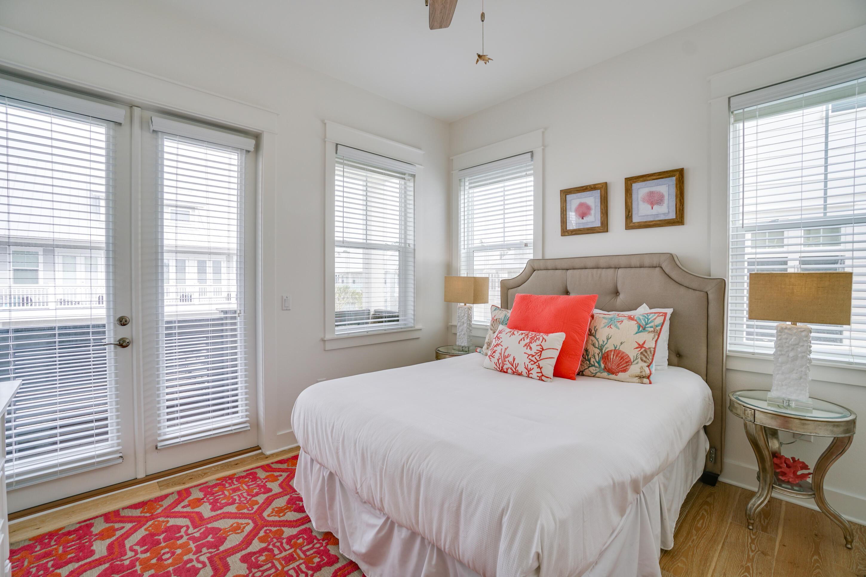 Bright bedroom with a white bed, coral accent pillows, seashell-patterned cushions, and a vivid coral rug.
