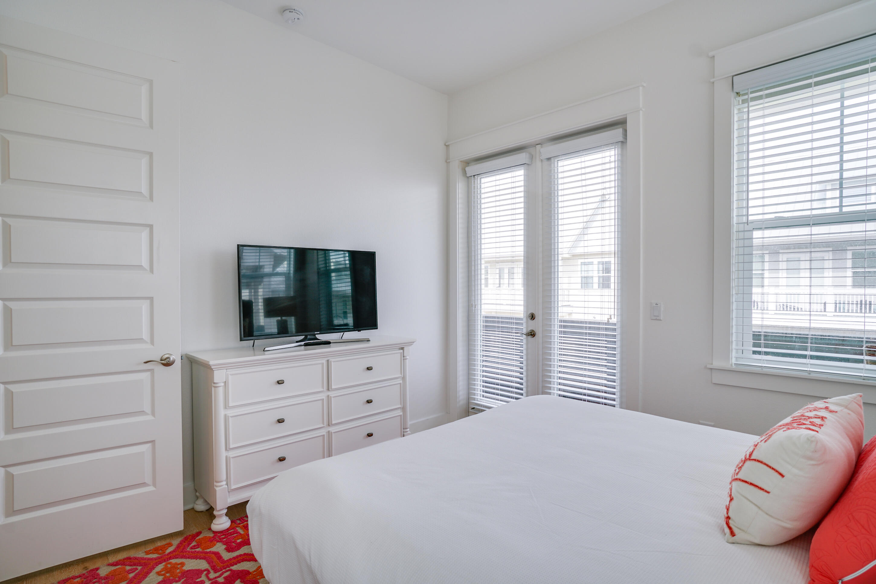 Bedroom with a white bed, red accent pillows, and a dresser with a television beside glass doors.