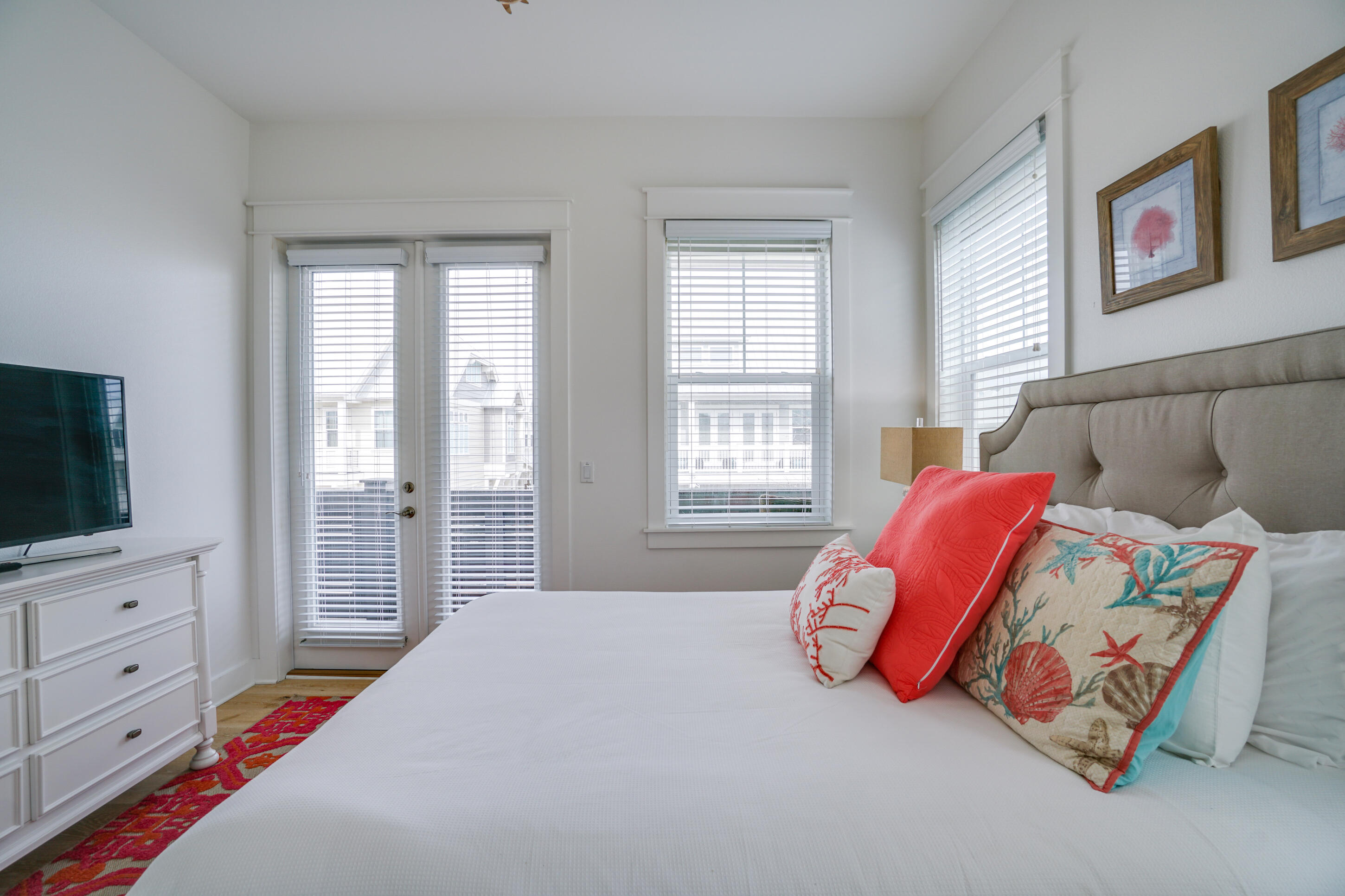 Furnished bedroom with a white bed, coral coastal pillows, gray headboard, and glass doors with blinds.