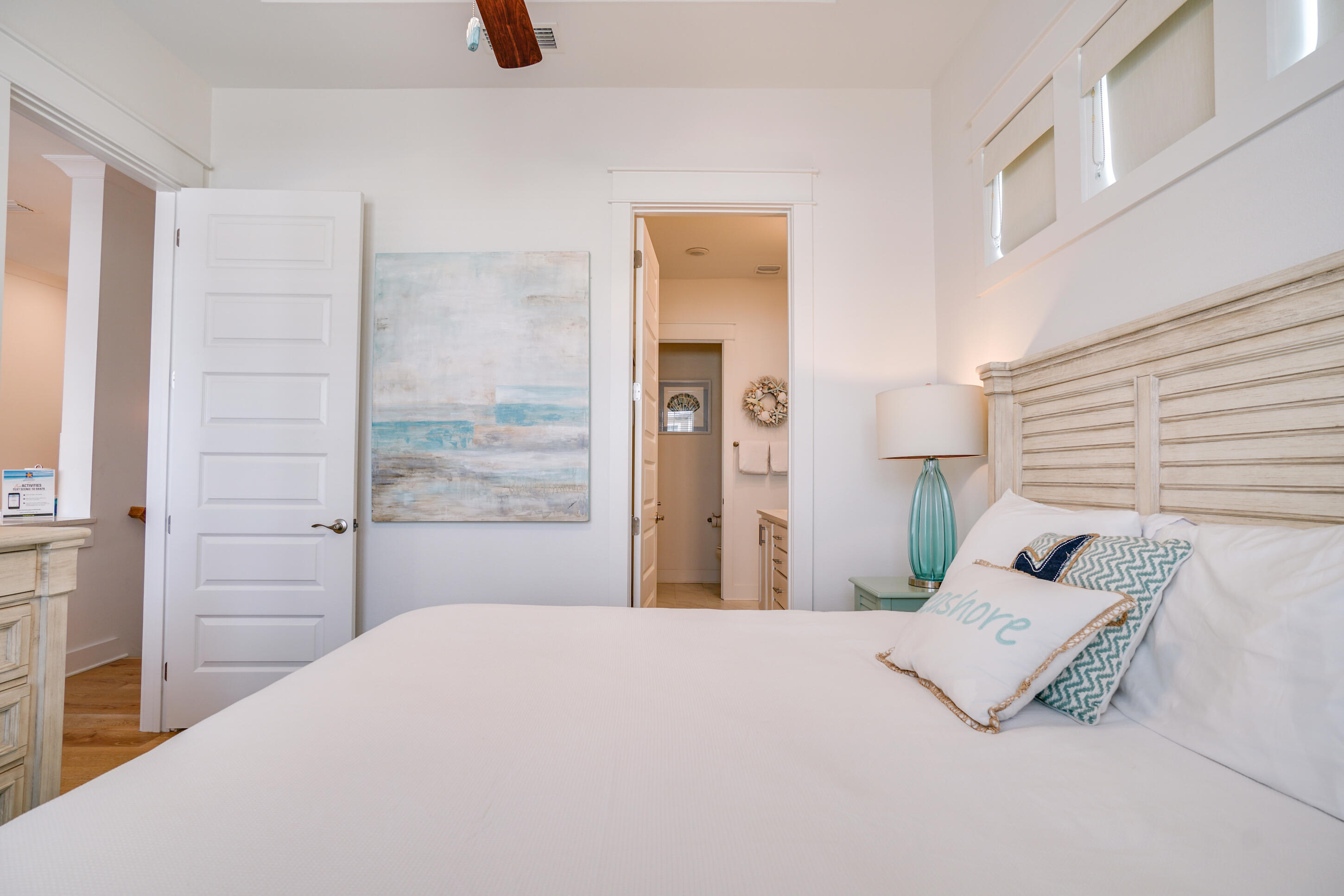 Bedroom with a white bed, weathered wood headboard, turquoise lamp, and open bathroom doorway.