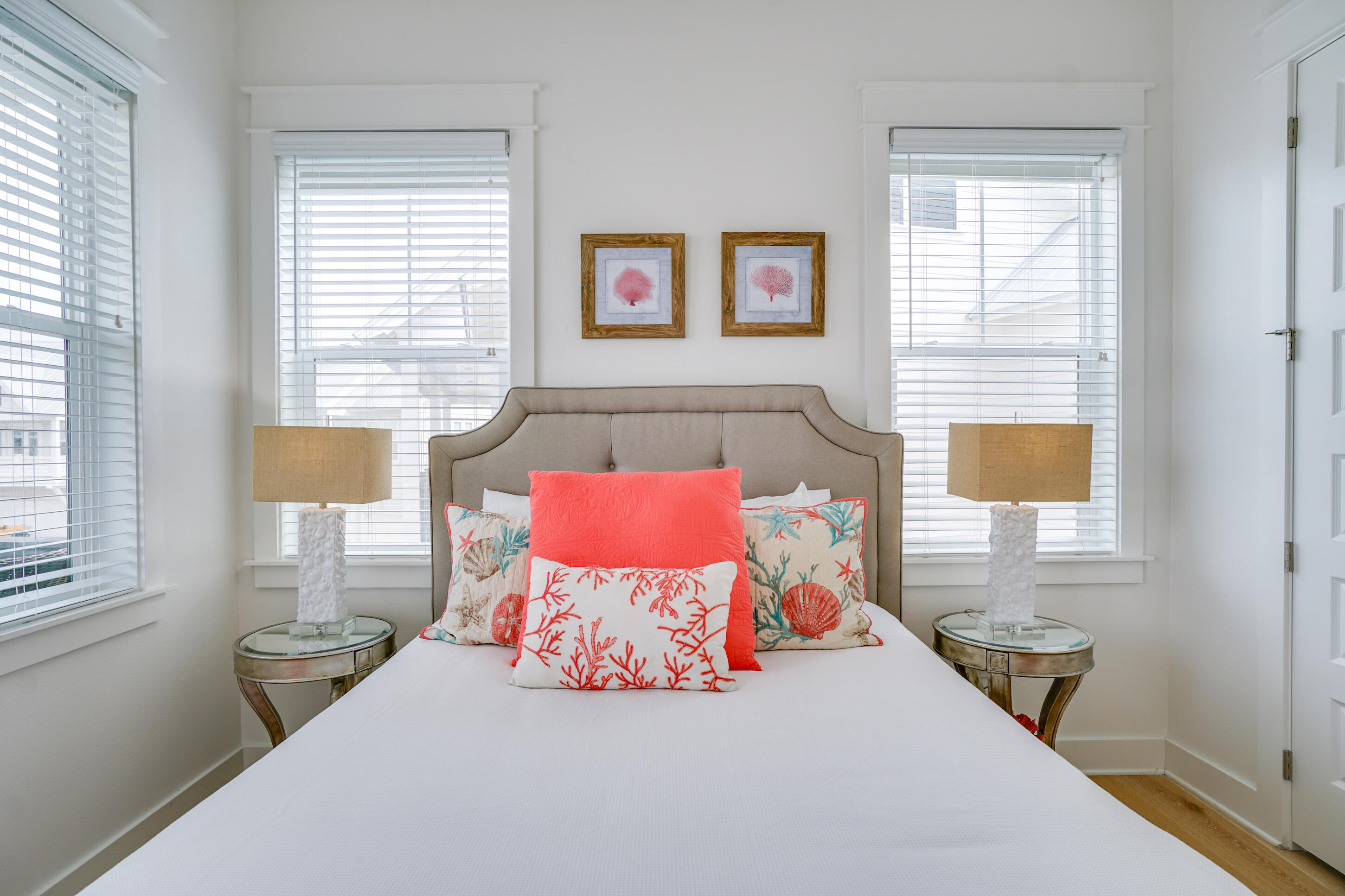 Bedroom with a gray upholstered headboard, coral pillows, and windows with white blinds.