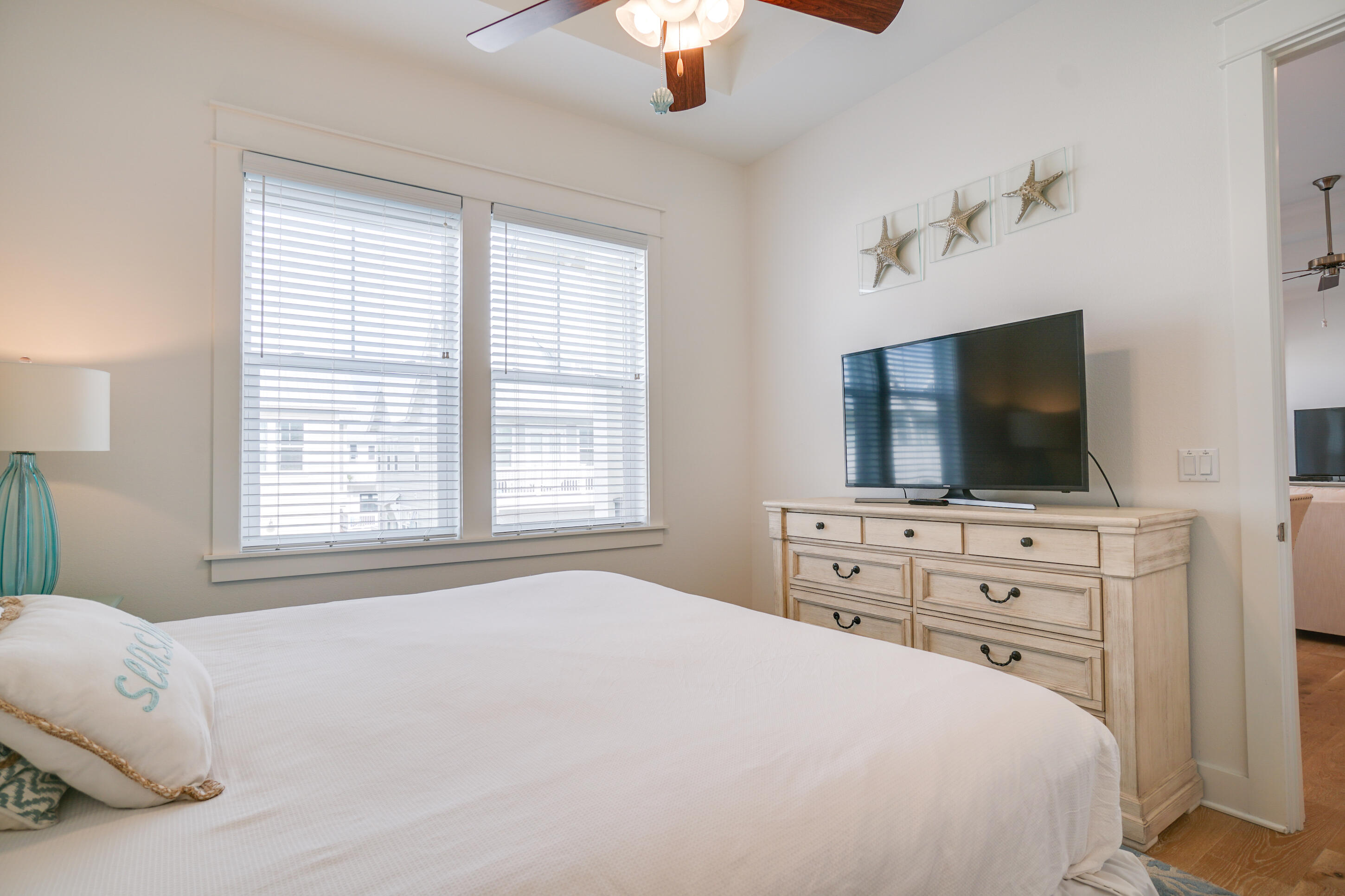 Staged bedroom with a white bed, starfish wall art, and a television on a light wood dresser.