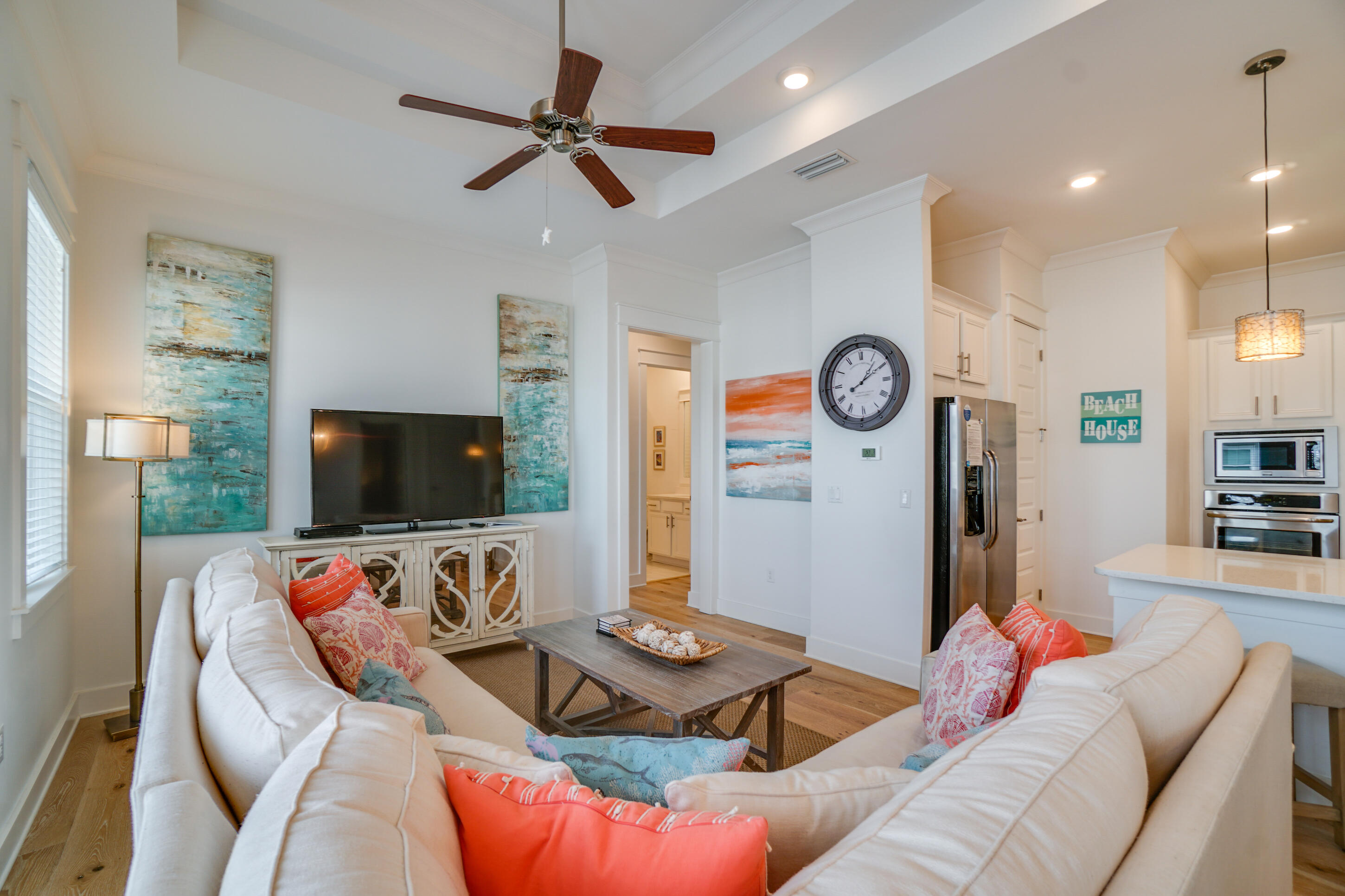 Open-plan living room with white sofas, colorful coastal cushions, and a kitchen with stainless steel appliances.