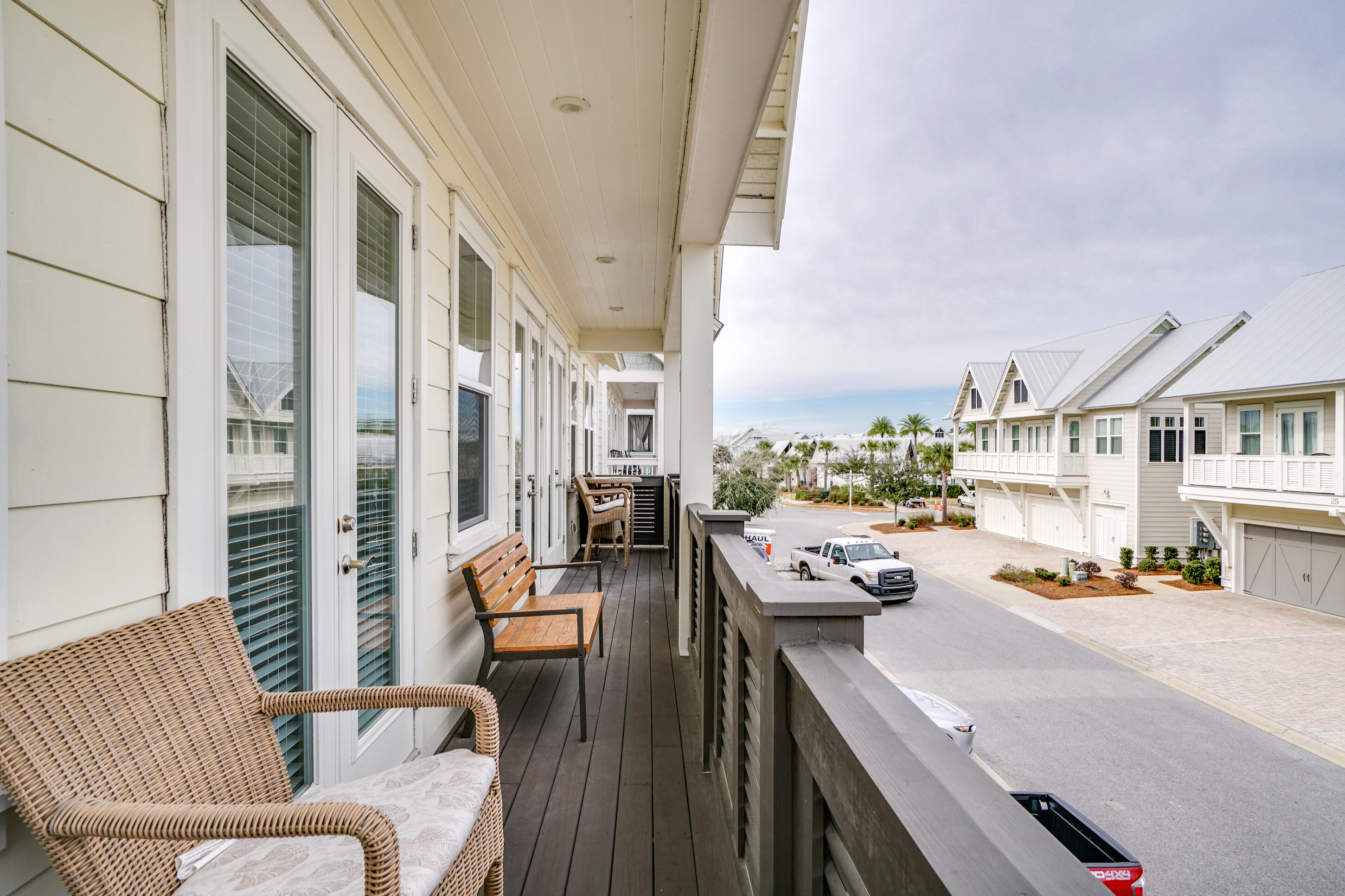 Covered balcony with wicker seating overlooking a street of light-colored homes with white metal roofs.
