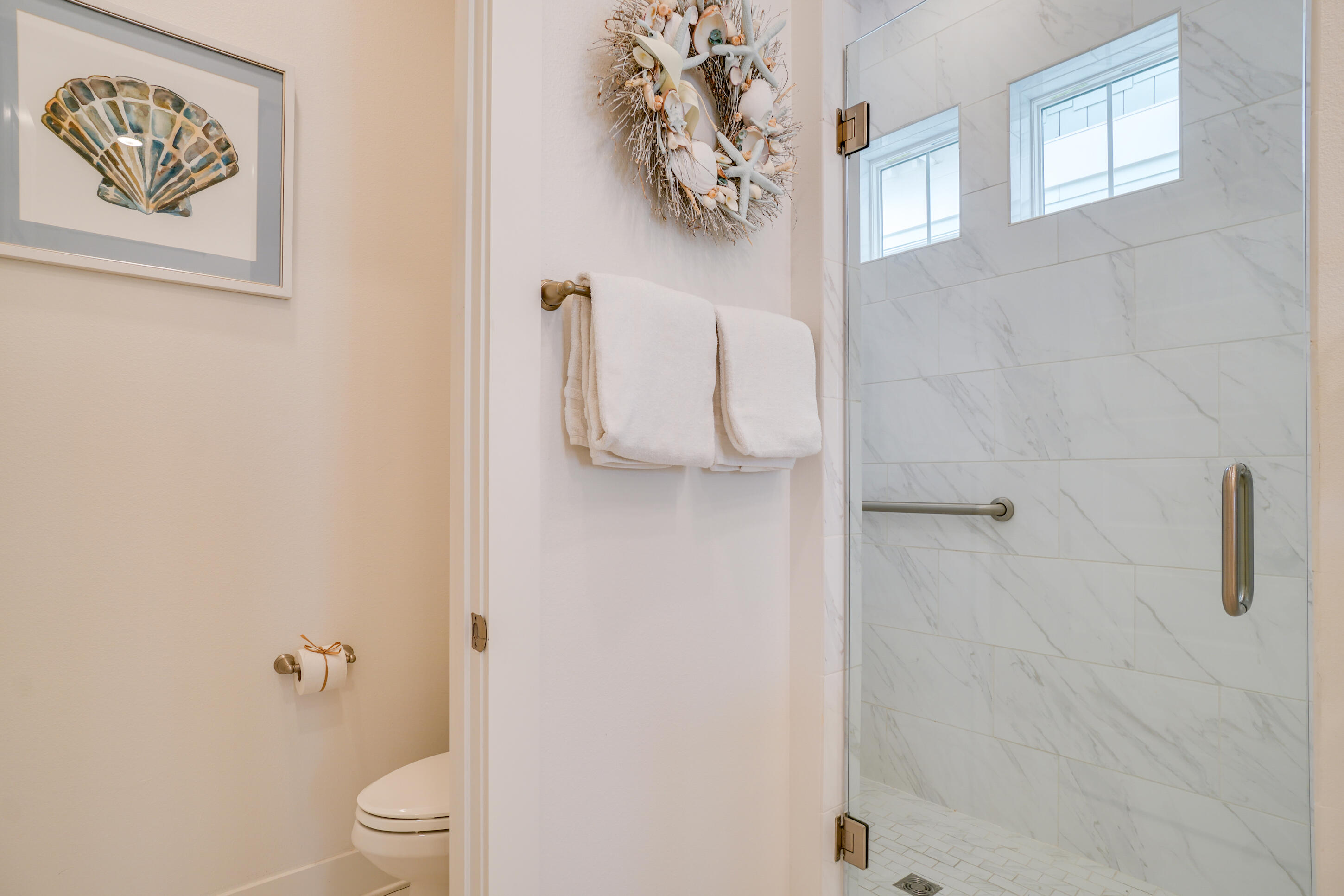 Bathroom with a marble-look tiled glass shower, high windows, and coastal shell décor.