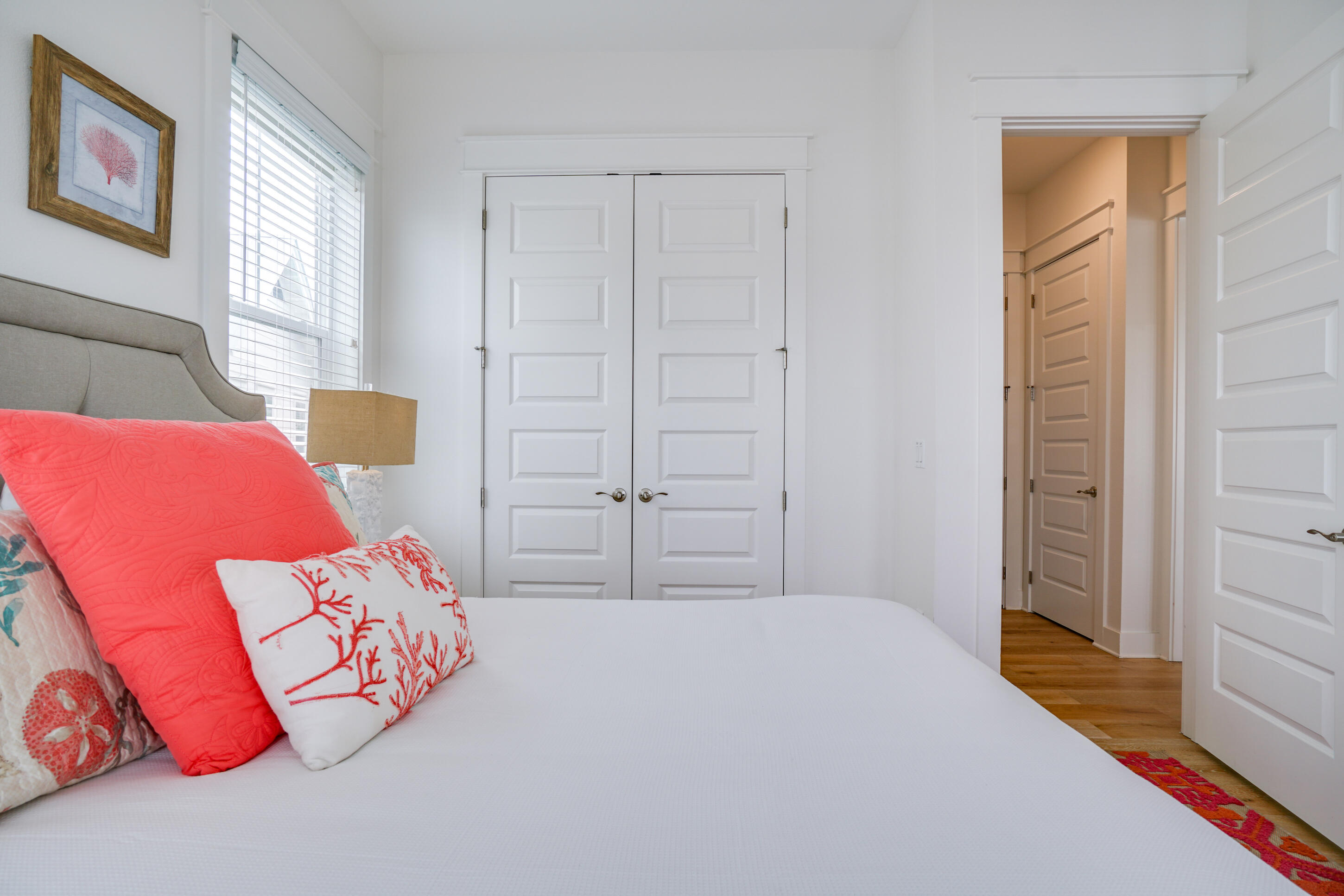 Bright bedroom with a white bed, coral pillows, paneled closet doors, and a window with blinds.