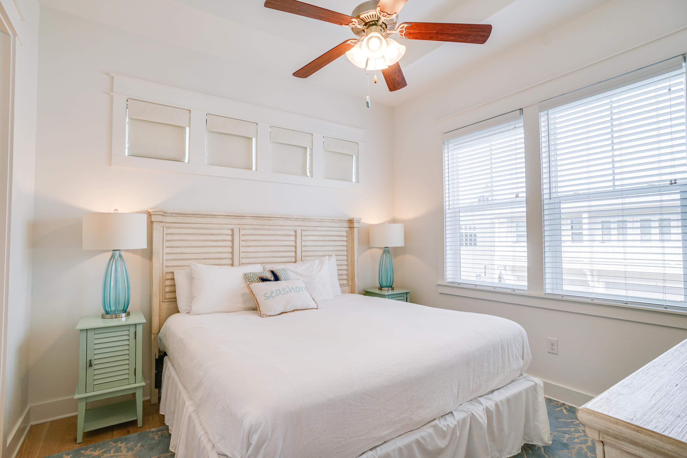 Bedroom with a white bed, pale wood headboard, turquoise lamps, and wide windows with blinds.