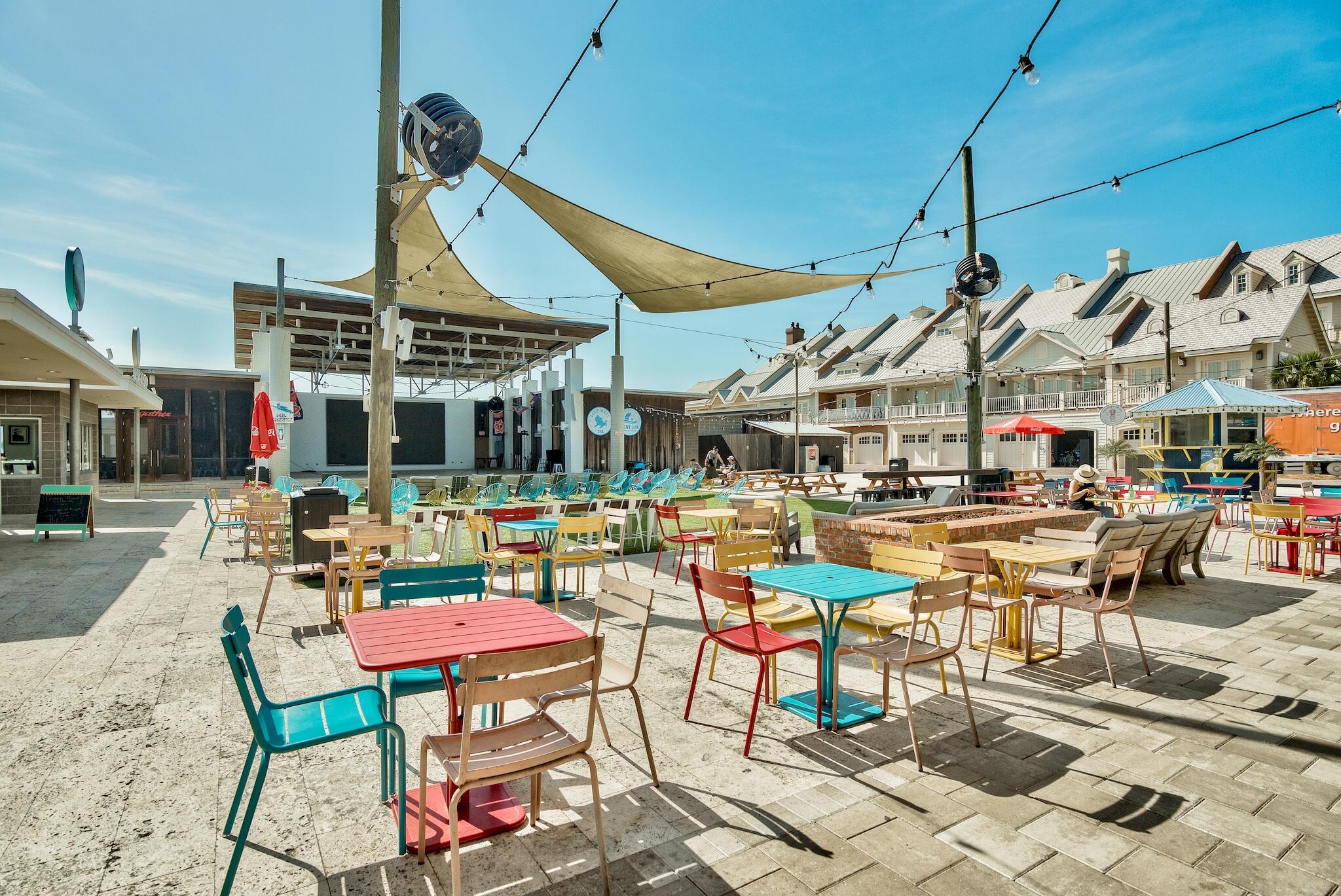 Outdoor courtyard with colorful café seating beneath shade sails and string lights, beside townhomes.