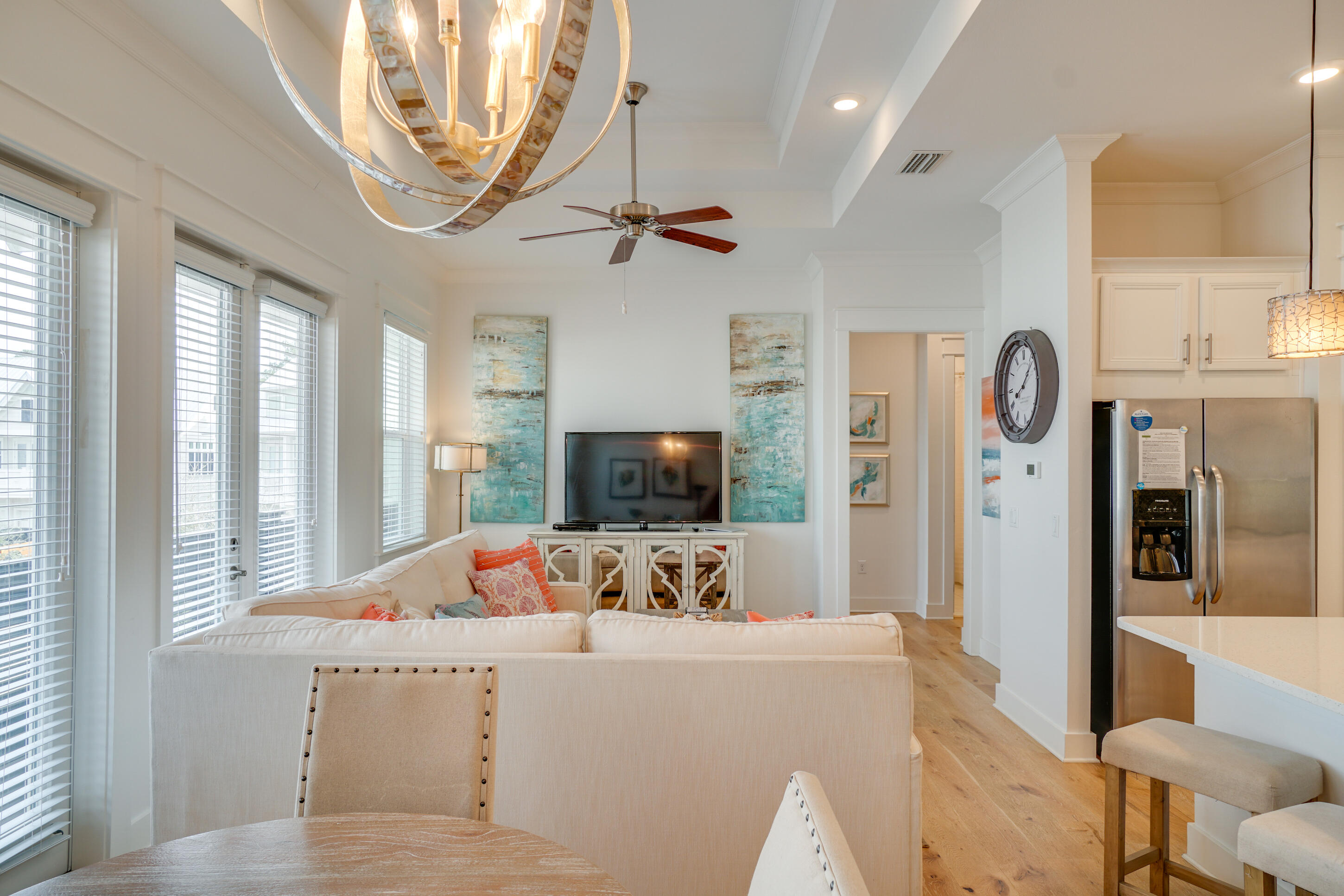 Open-plan living room and kitchen with tall windows, a cream sectional, pale wood floors, and stainless steel refrigerator.