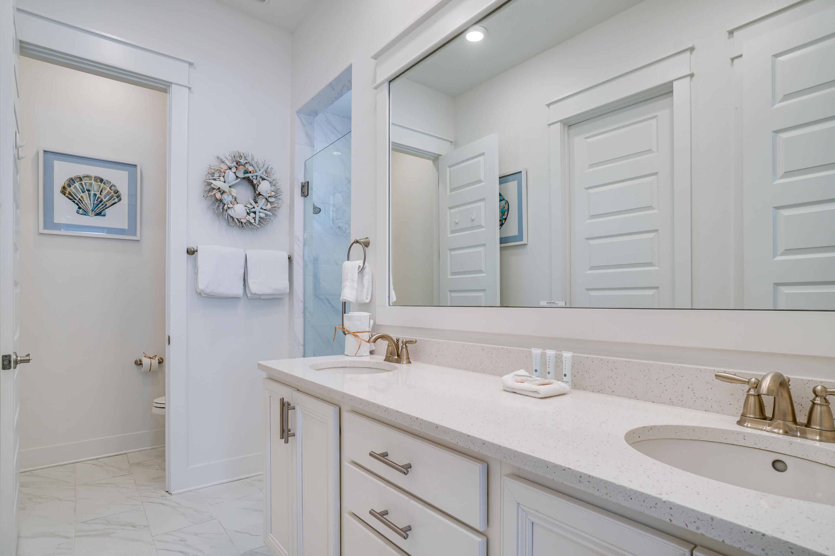Bright bathroom with a white double-sink vanity, brass-tone faucets, and a marble-look tiled shower.