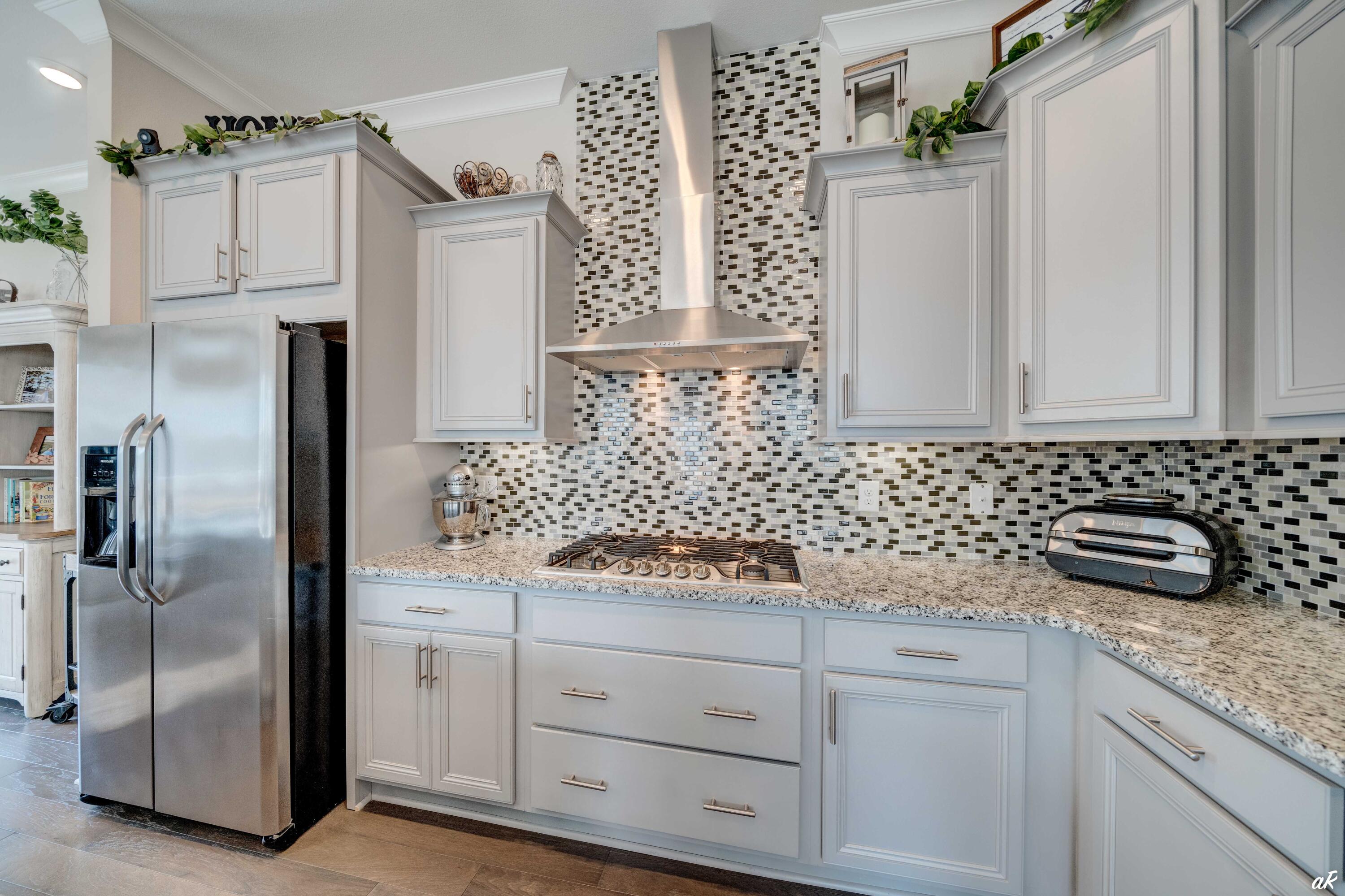 Kitchen with white cabinets, granite counters, mosaic backsplash, stainless refrigerator, and gas cooktop.