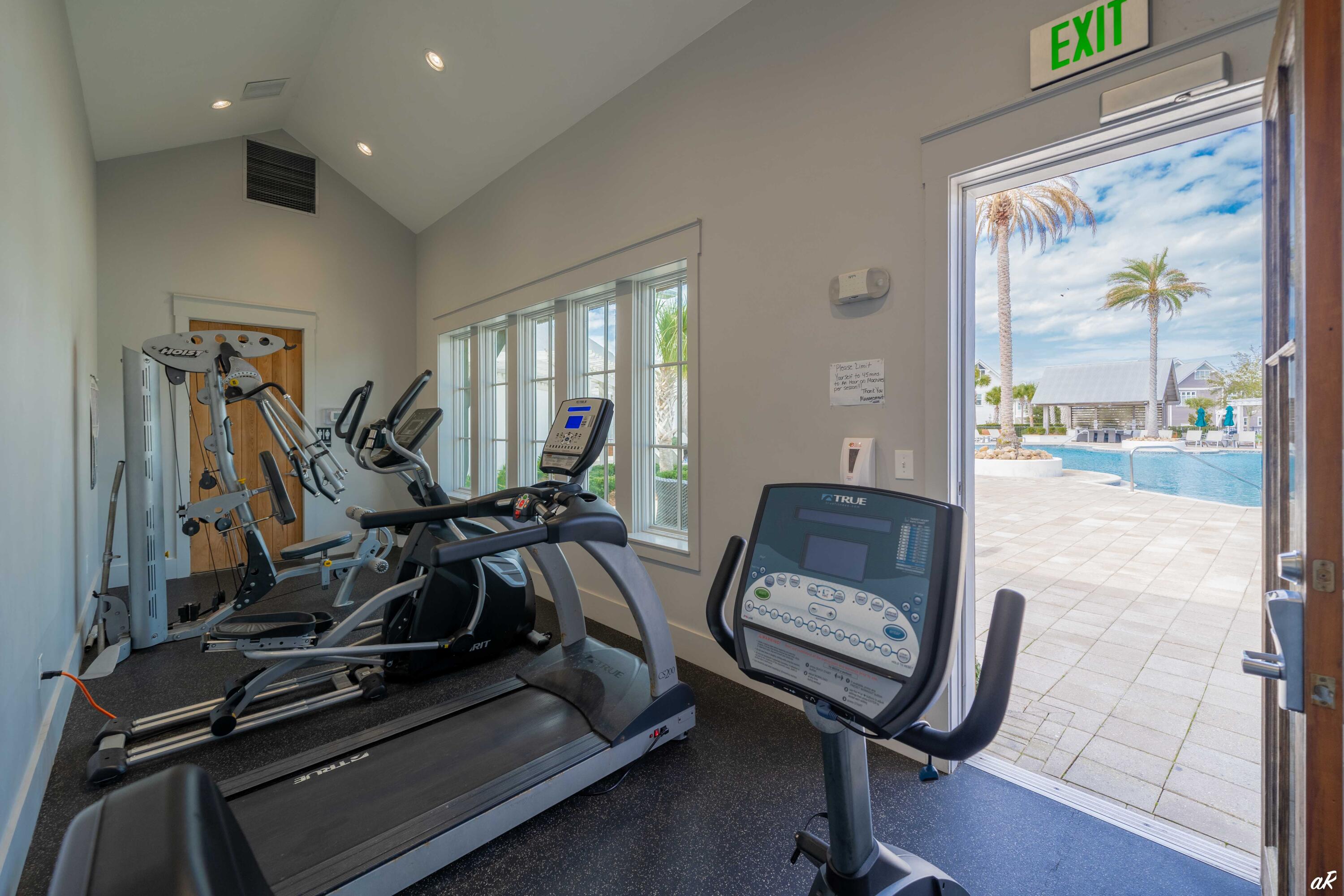 Fitness room with exercise machines and an open doorway overlooking a pool and palm trees.