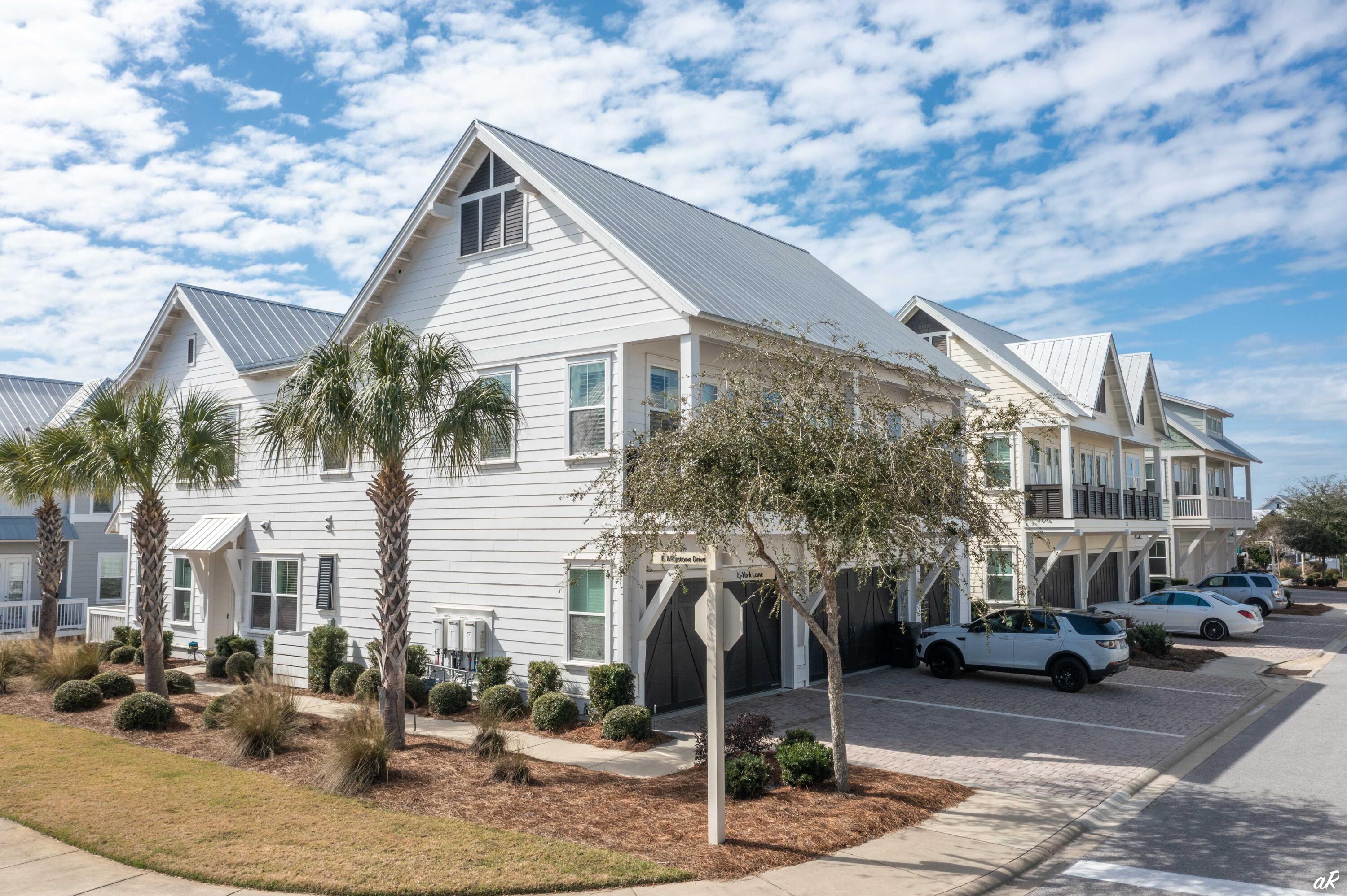 White coastal townhome with a steep metal roof, palm trees, and paved parking.