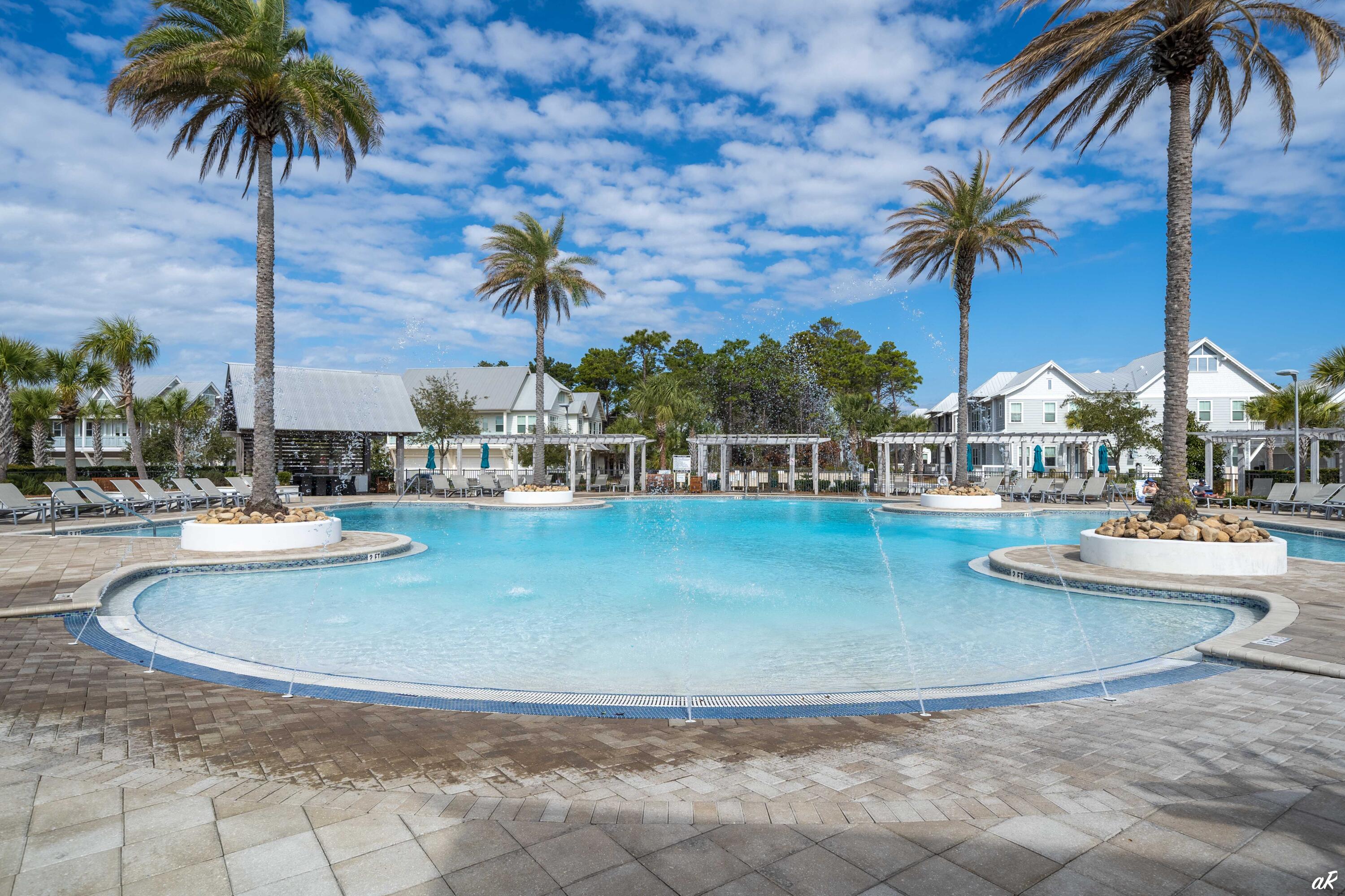 Community swimming pool with palm trees, fountains, lounge chairs, and white coastal homes.