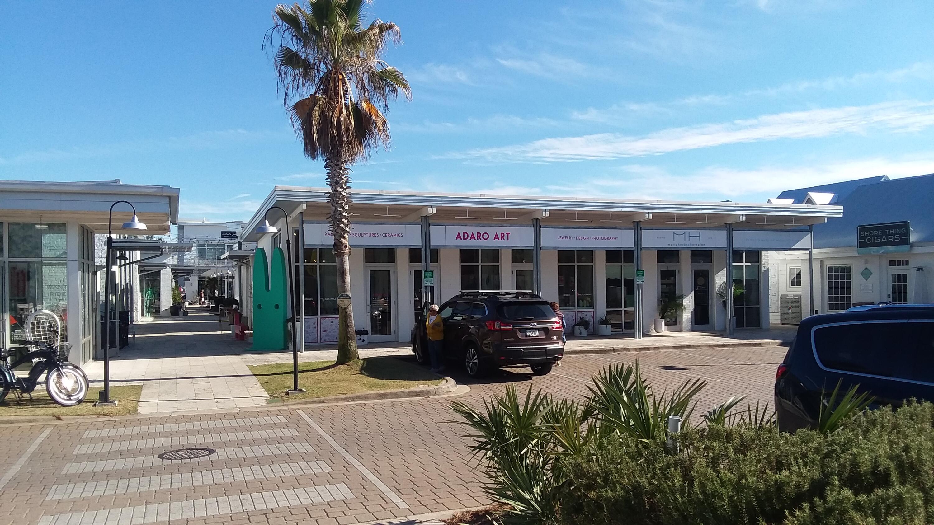 Retail storefronts beneath a long canopy, with a palm tree and brick-paved parking.