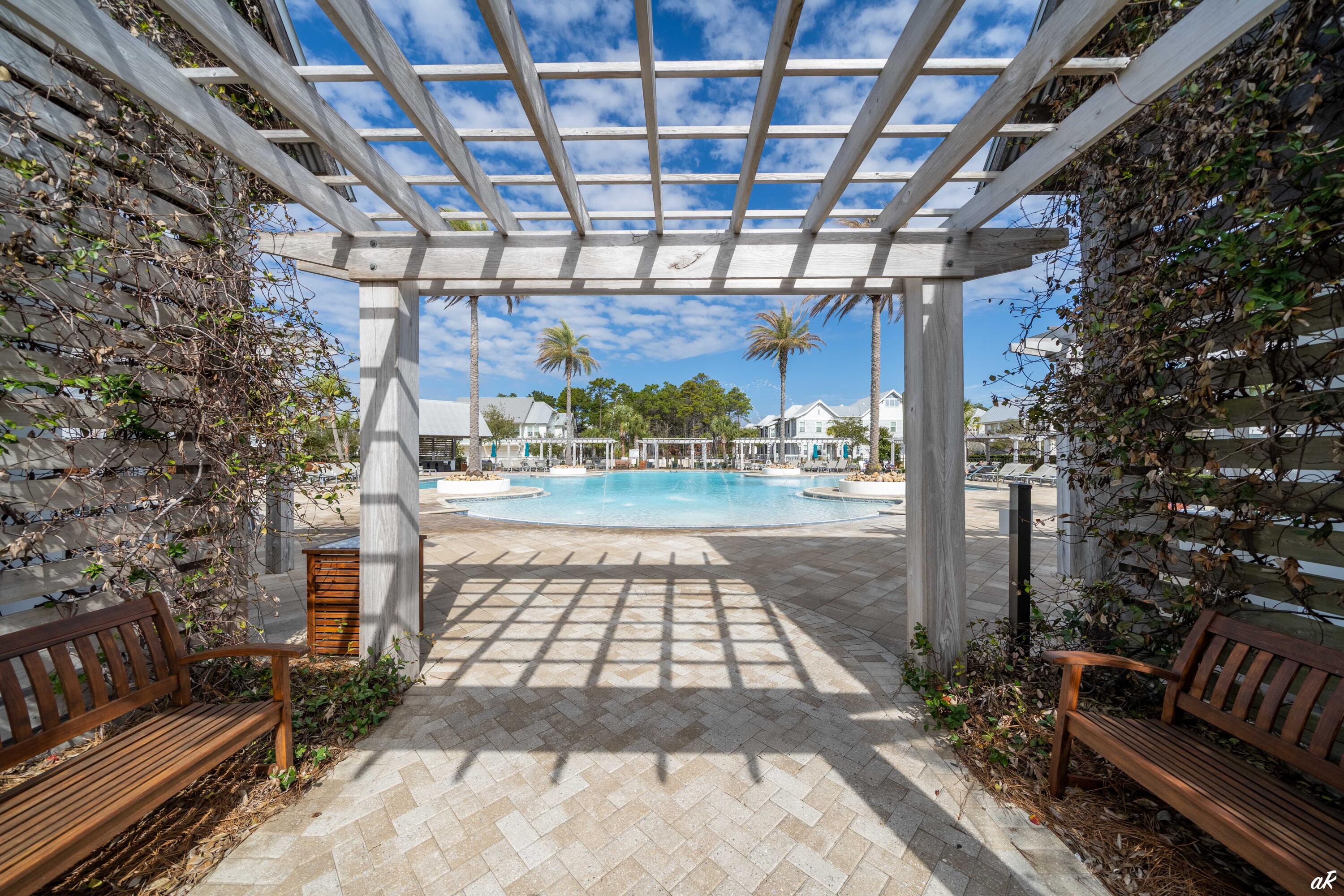 Community pool framed by a vine-covered pergola, with palm trees and homes beyond.