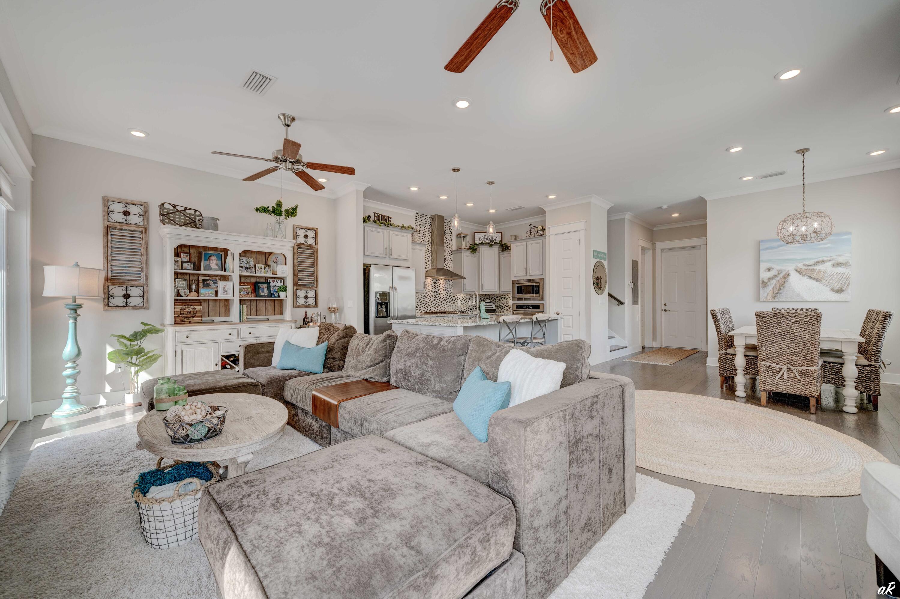 Open-plan living room with a large gray sectional, coastal decor, and adjoining white kitchen and dining area.