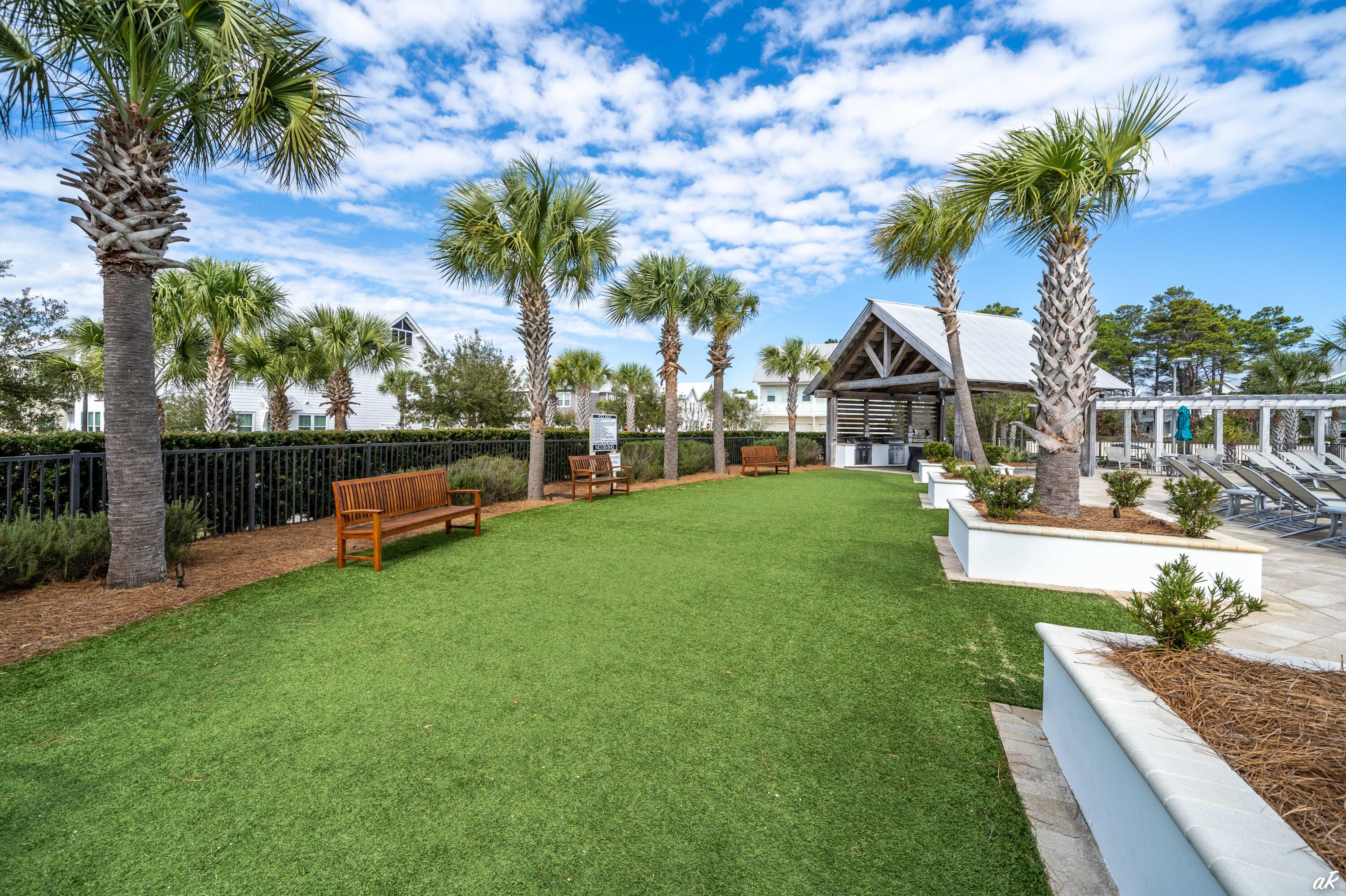 Landscaped community lawn with palm trees, wooden benches, a covered pavilion, and poolside lounge chairs.