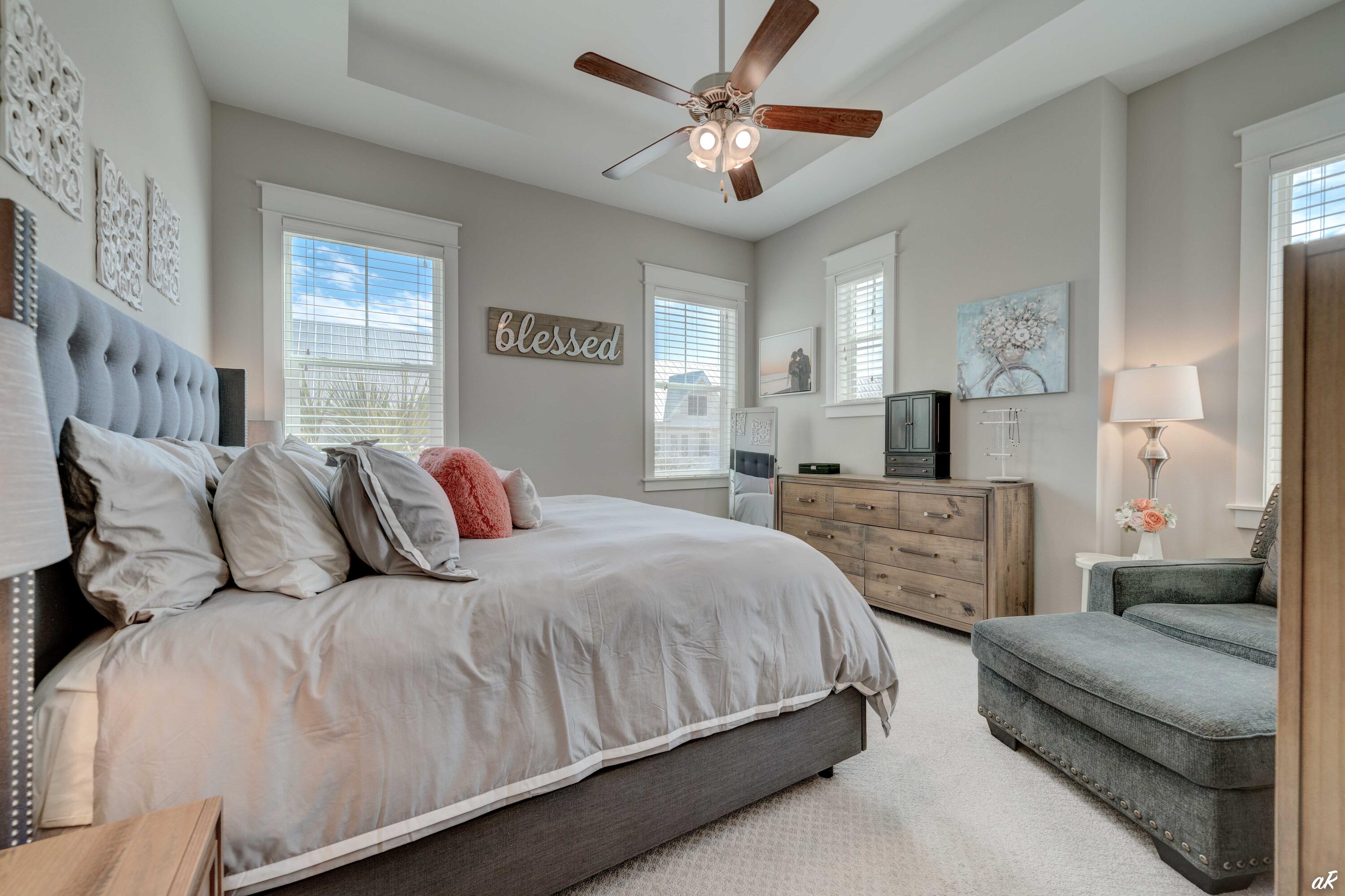 Furnished bedroom with a large gray bed, several windows, a wooden dresser, and an upholstered armchair.