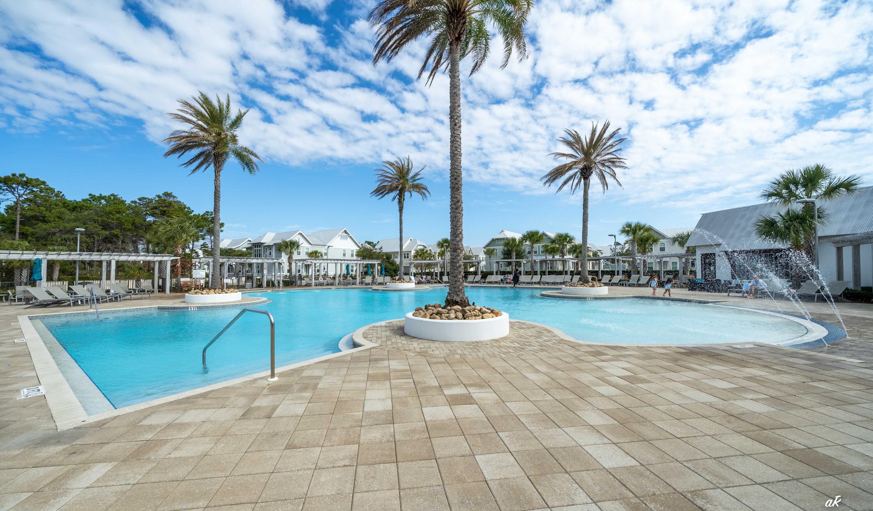Outdoor pool with palm-tree planters, a shallow splash area, and lounge seating beside coastal homes.