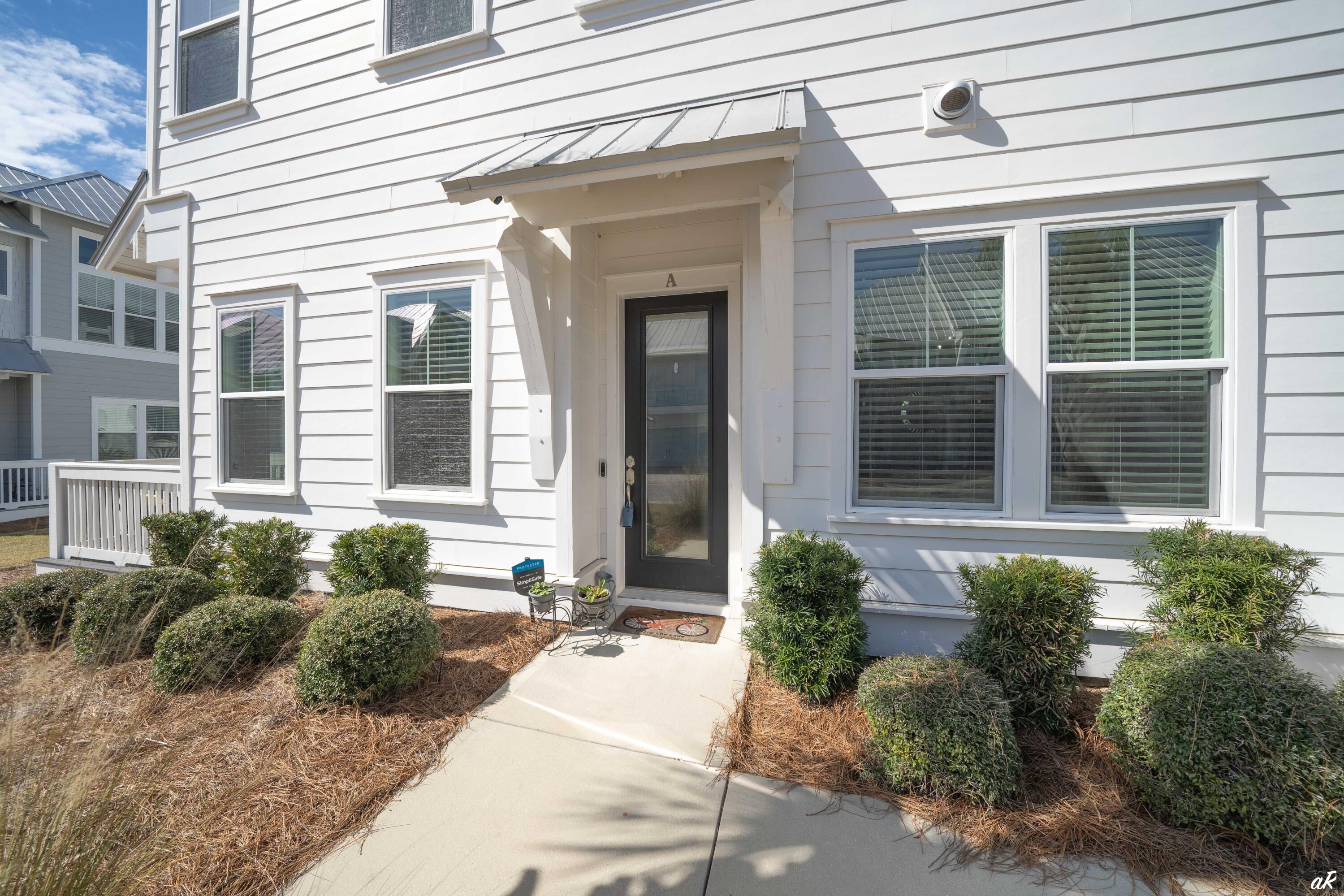 White-sided home entrance with a black glass-paneled door, metal awning, and landscaped walkway.