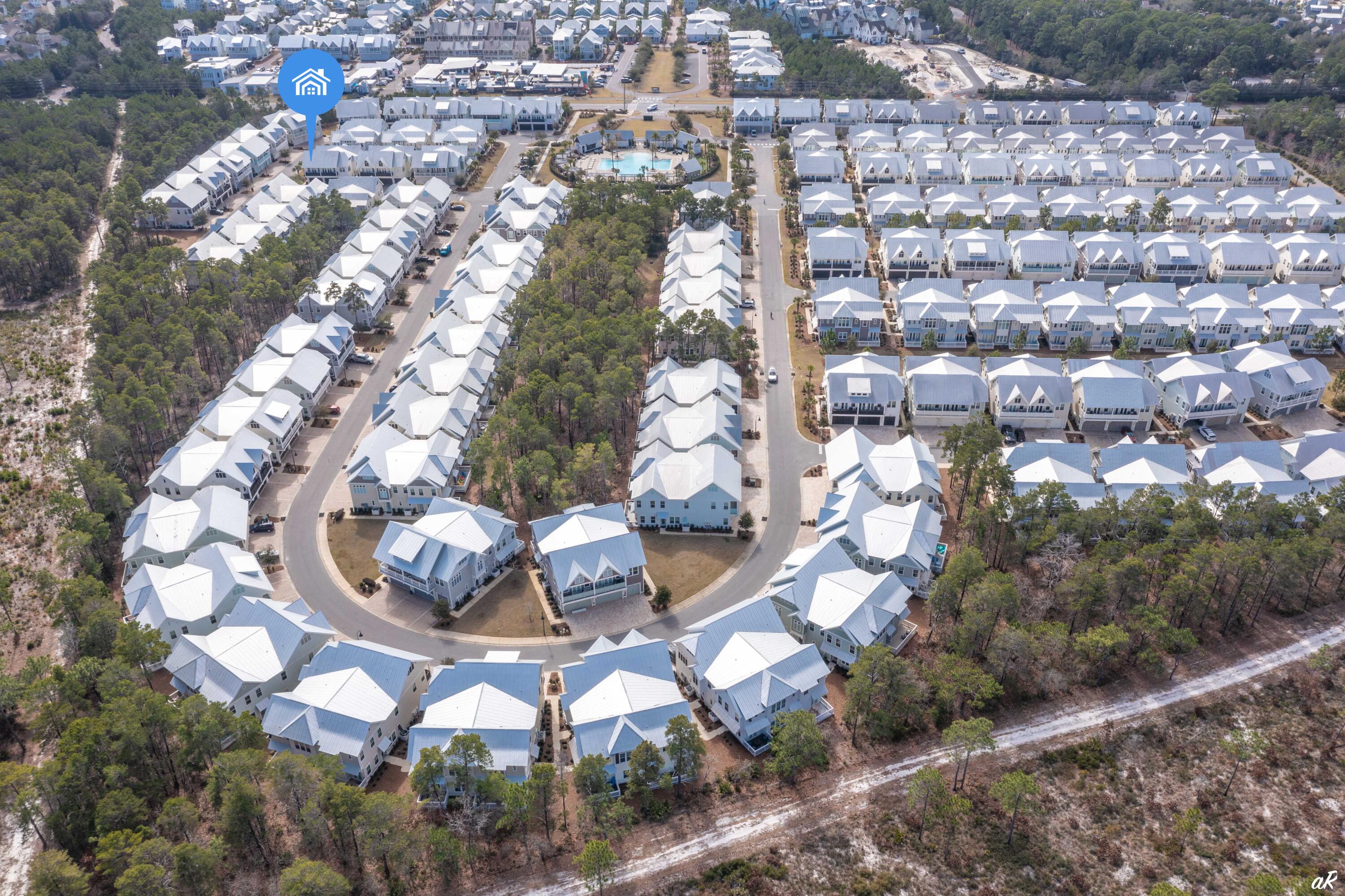Aerial view of a residential neighborhood with white-roofed homes, a central pool, and surrounding woodland.