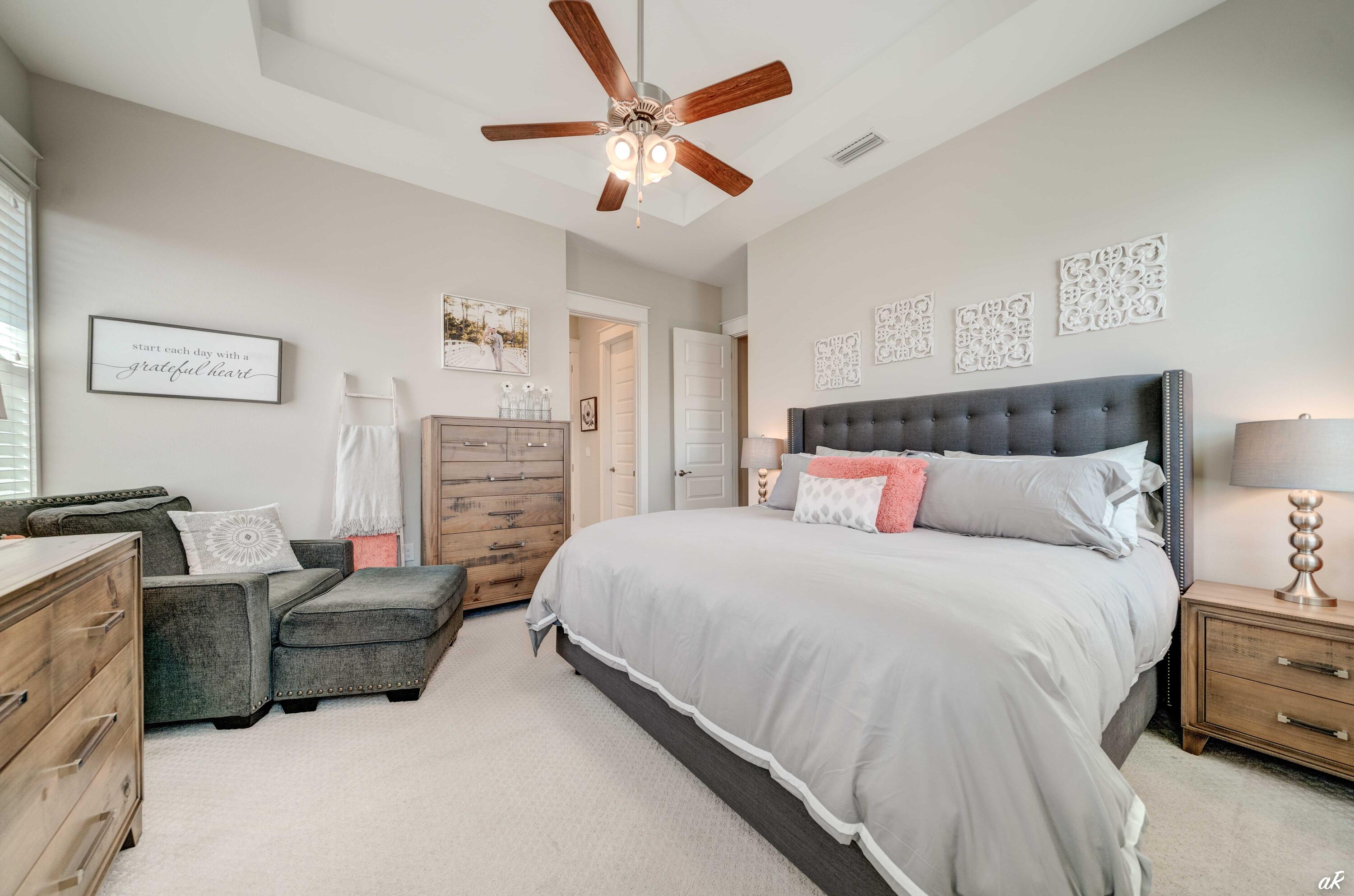 Furnished bedroom with a gray tufted bed, wood dressers, and a sitting area.
