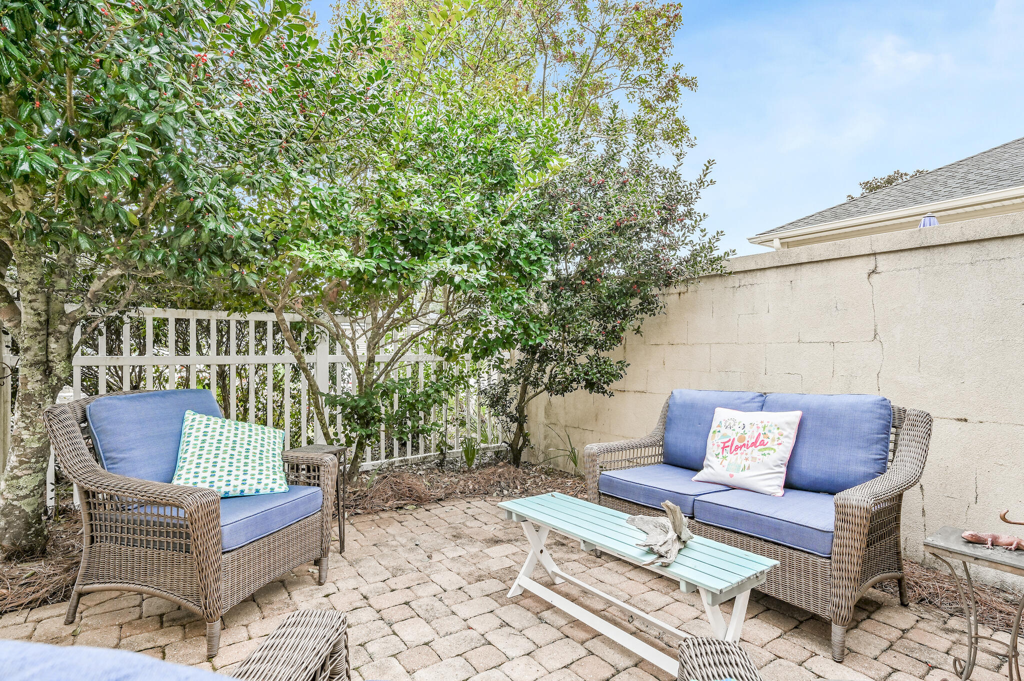 Brick-paved patio with blue-cushioned wicker seating, a pale aqua table, and leafy trees.