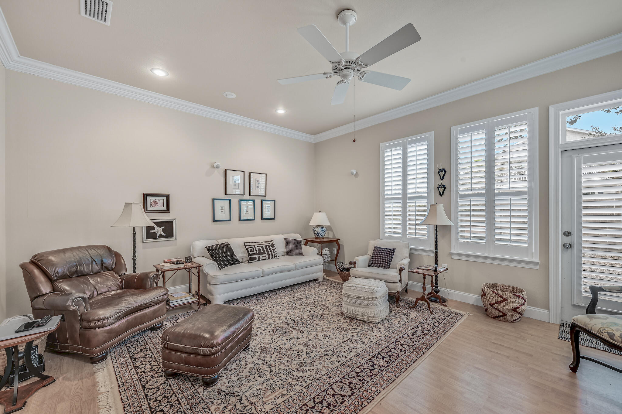 Living room with a white sofa, leather armchair, patterned rug, and tall windows with plantation shutters.