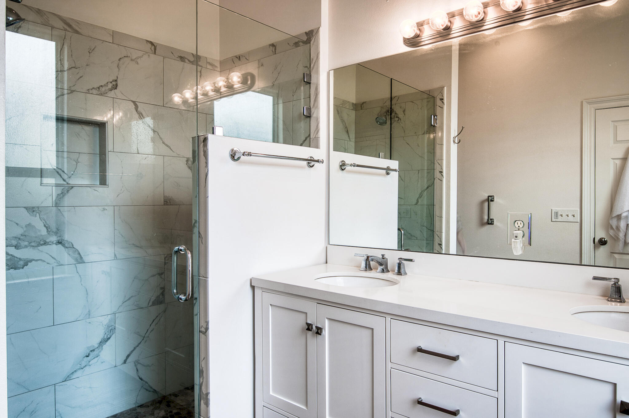 Bathroom with a glass shower, gray-veined tile, and a long double-sink vanity.