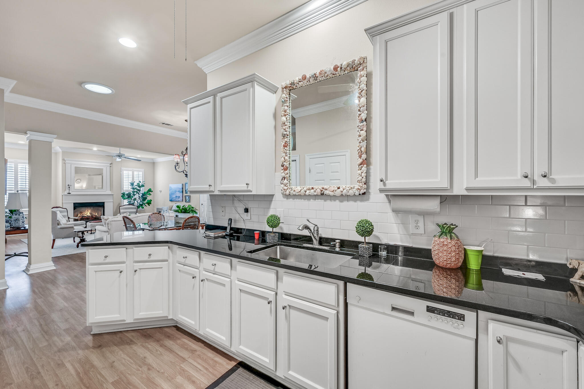 Kitchen with white cabinets, black countertops, and a shell-framed mirror, opening onto a living room with a fireplace.