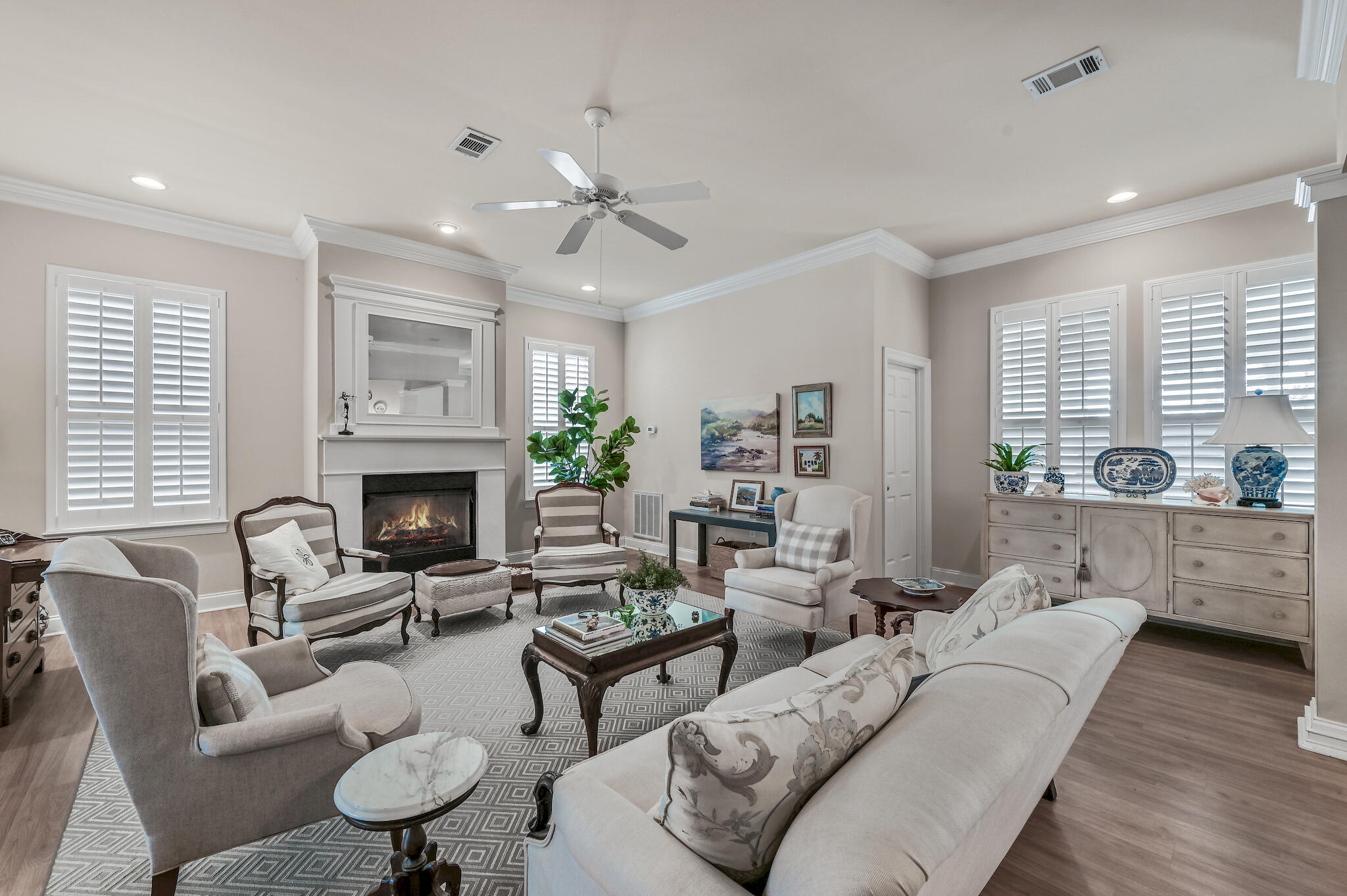 Living room with a white fireplace, shuttered windows, and pale seating around a patterned rug.