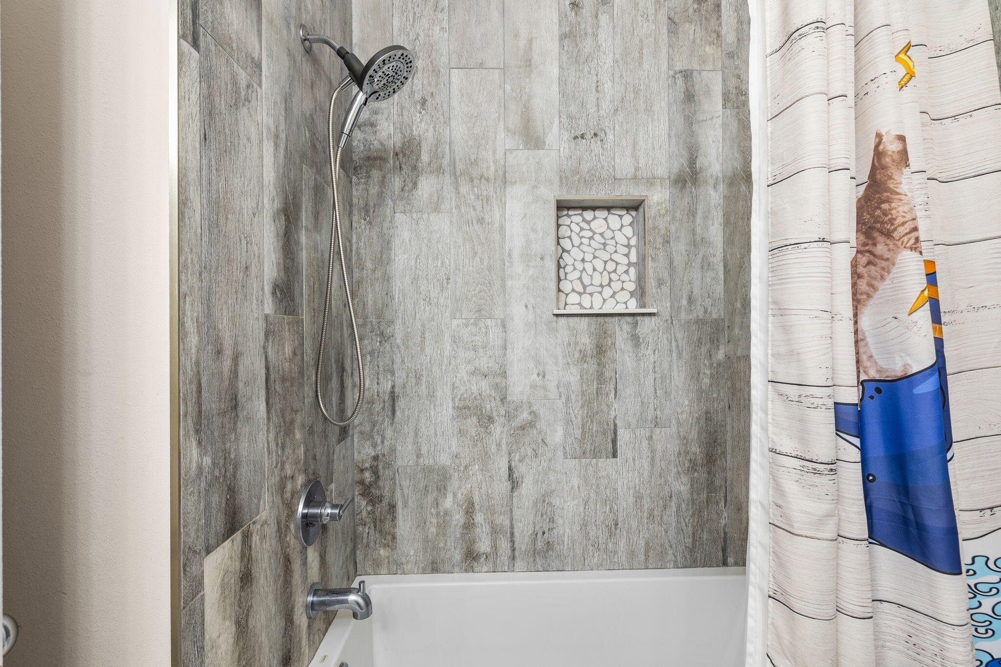 Bathroom tub and shower with gray tile, a pebble-lined niche, and a blue-and-white shower curtain.